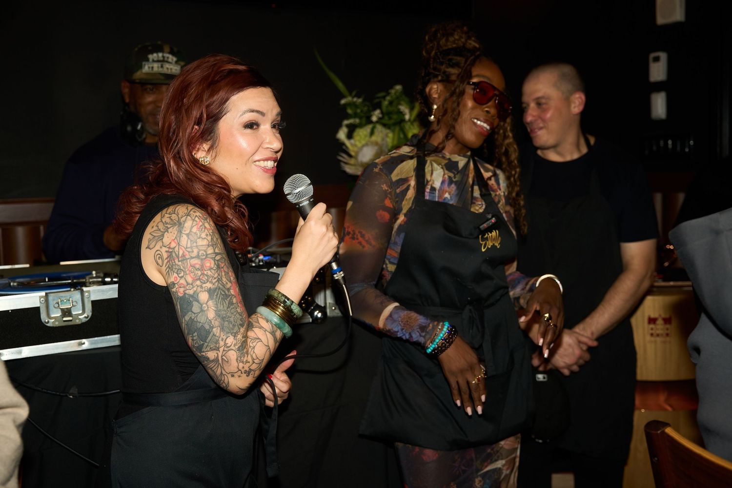 Tattooed woman with red hair speaks into a microphone while a woman in sunglasses and a man in black listen at an indoor event.