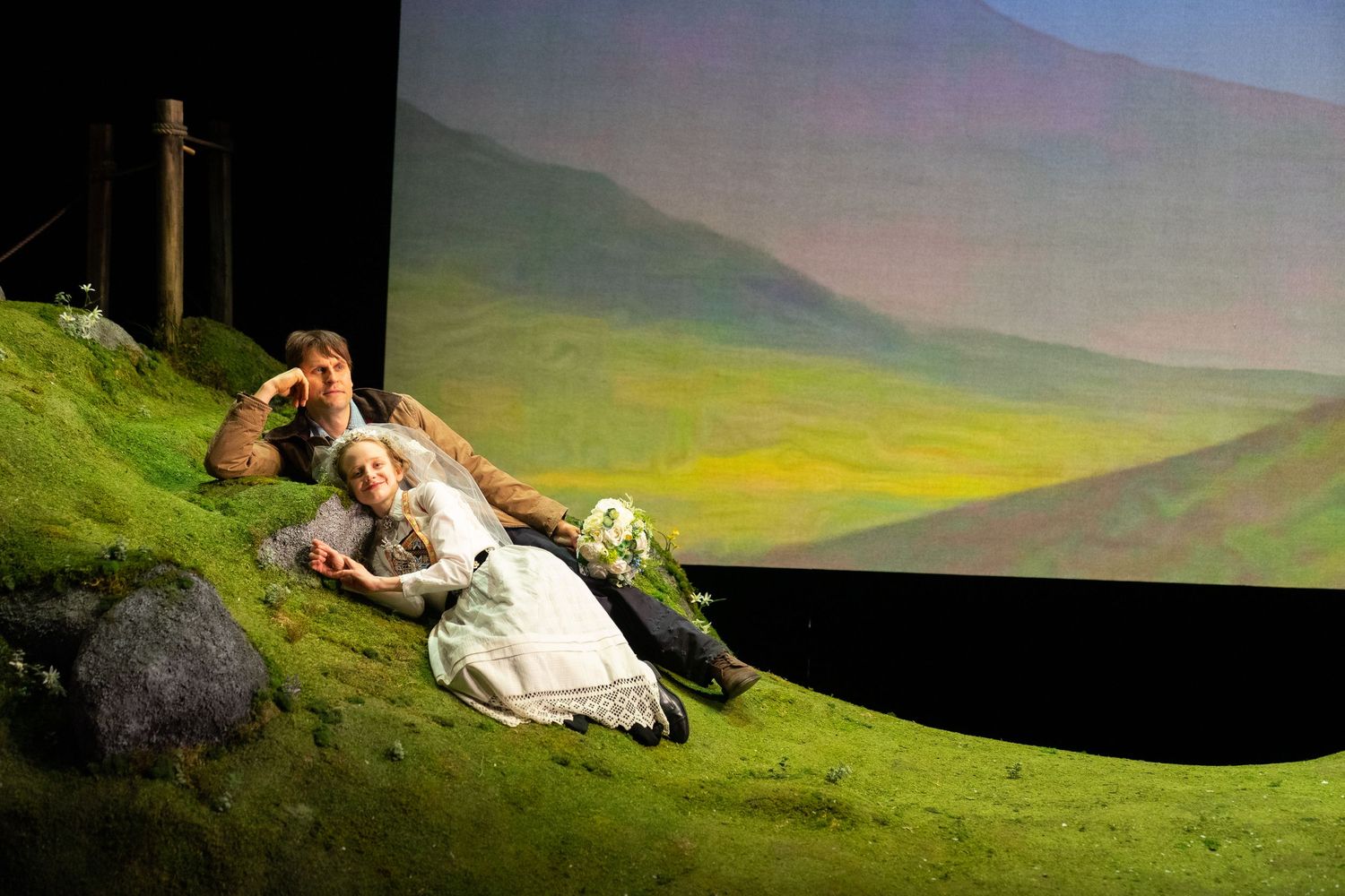 A couple rests together on a scenic hillside overlooking a misty mountain landscape at sunset.