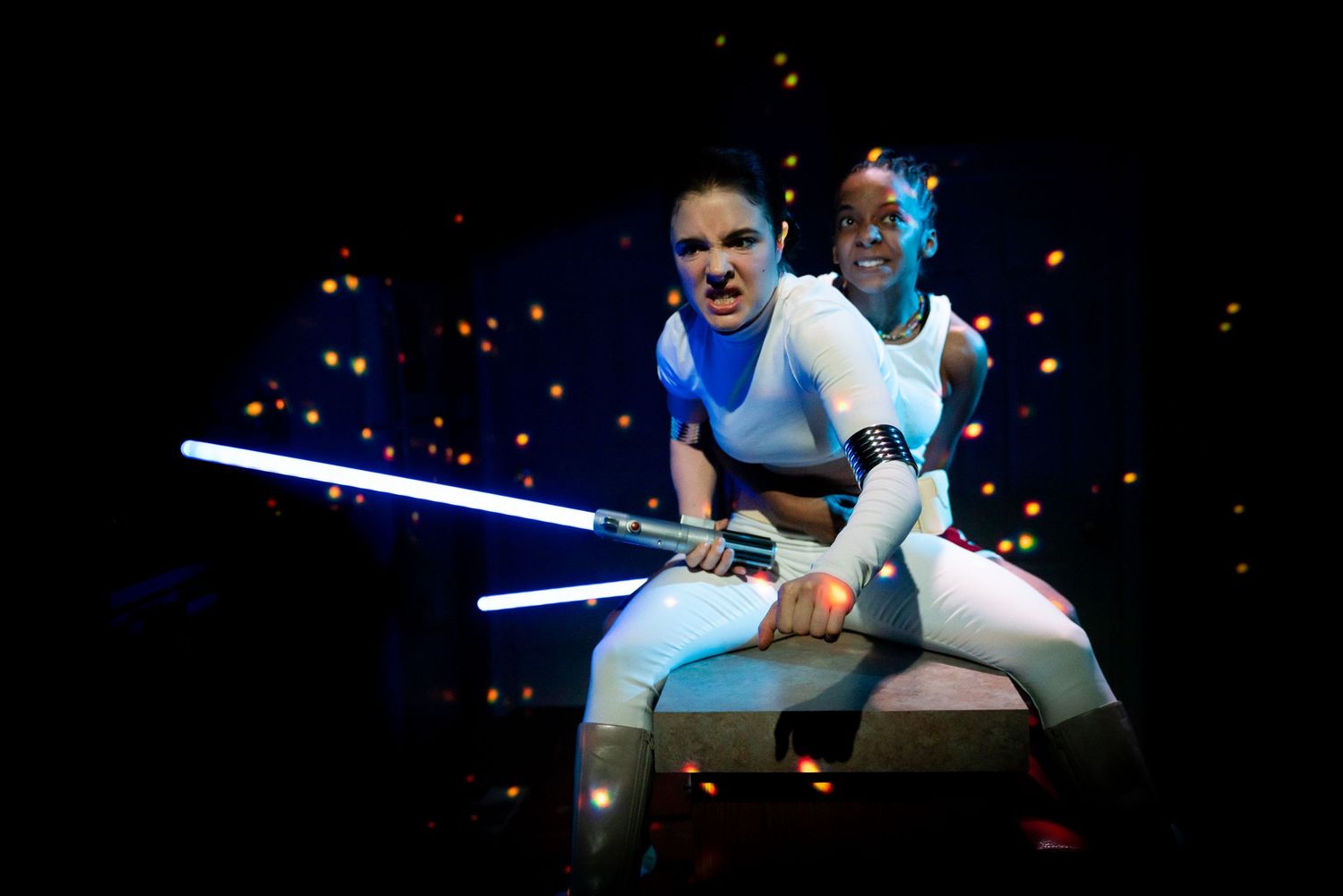 A person in white and light blue costume wields a glowing lightsaber while performing on stage with dark starry backdrop.