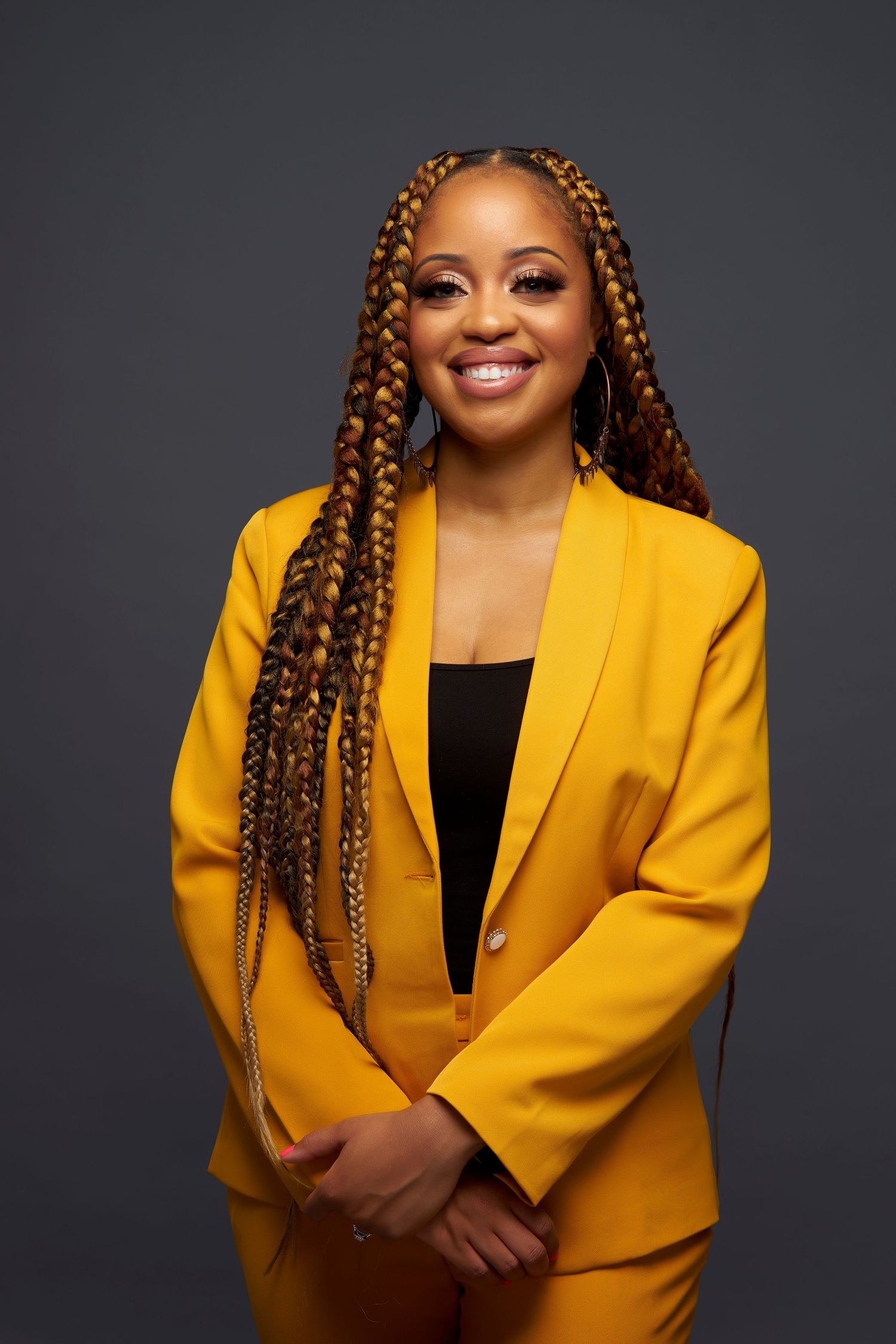 Professional studio portrait of someone wearing a vibrant yellow blazer with long braided hair against a dark background.