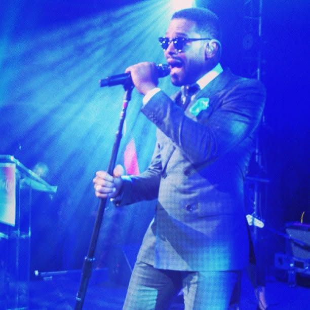 Male singer in sunglasses and a gray double-breasted suit performs into a microphone on stage under vibrant blue concert lighting.