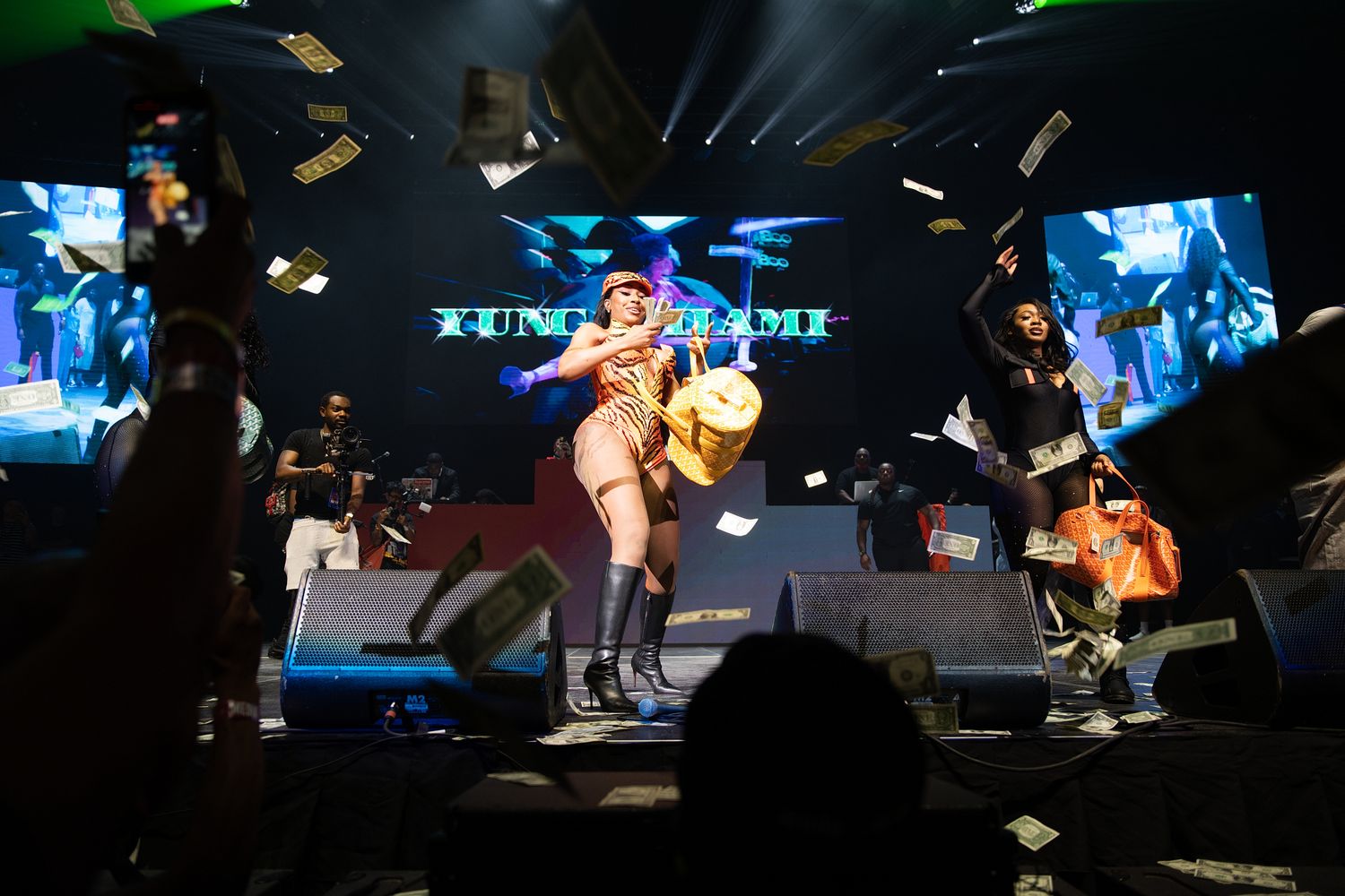 Yung Miami performs energetically on stage with dancers, money flying through the air, and vibrant LED backdrop.
