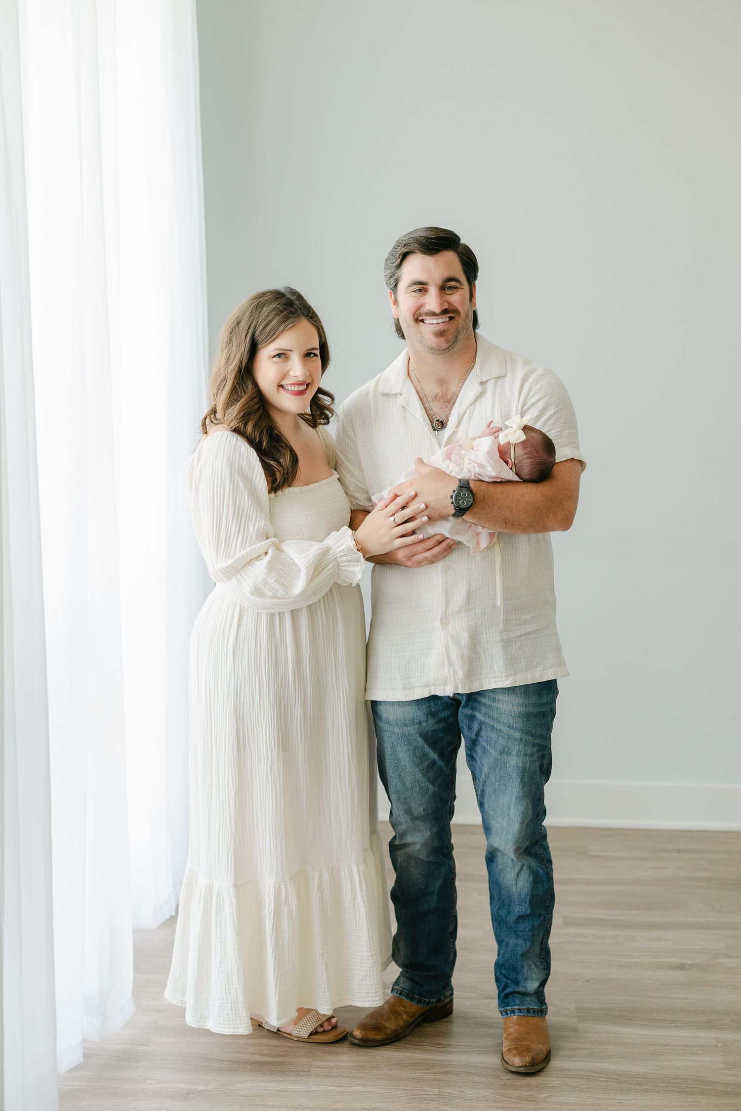 Family portrait with parents in white outfits holding newborn baby in bright studio setting.