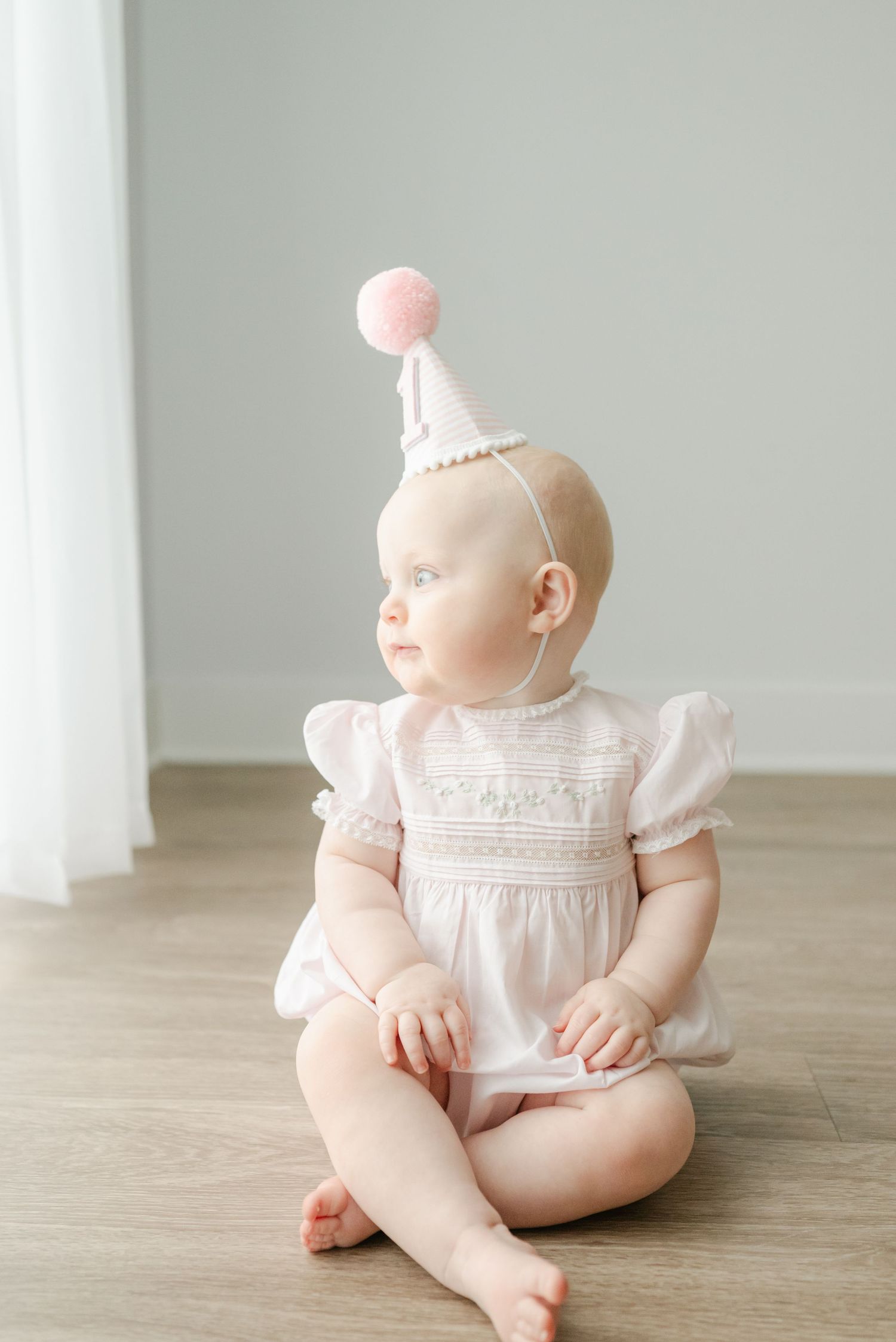 A baby wearing a white lace dress sits on a light wooden floor in a minimalist setting.