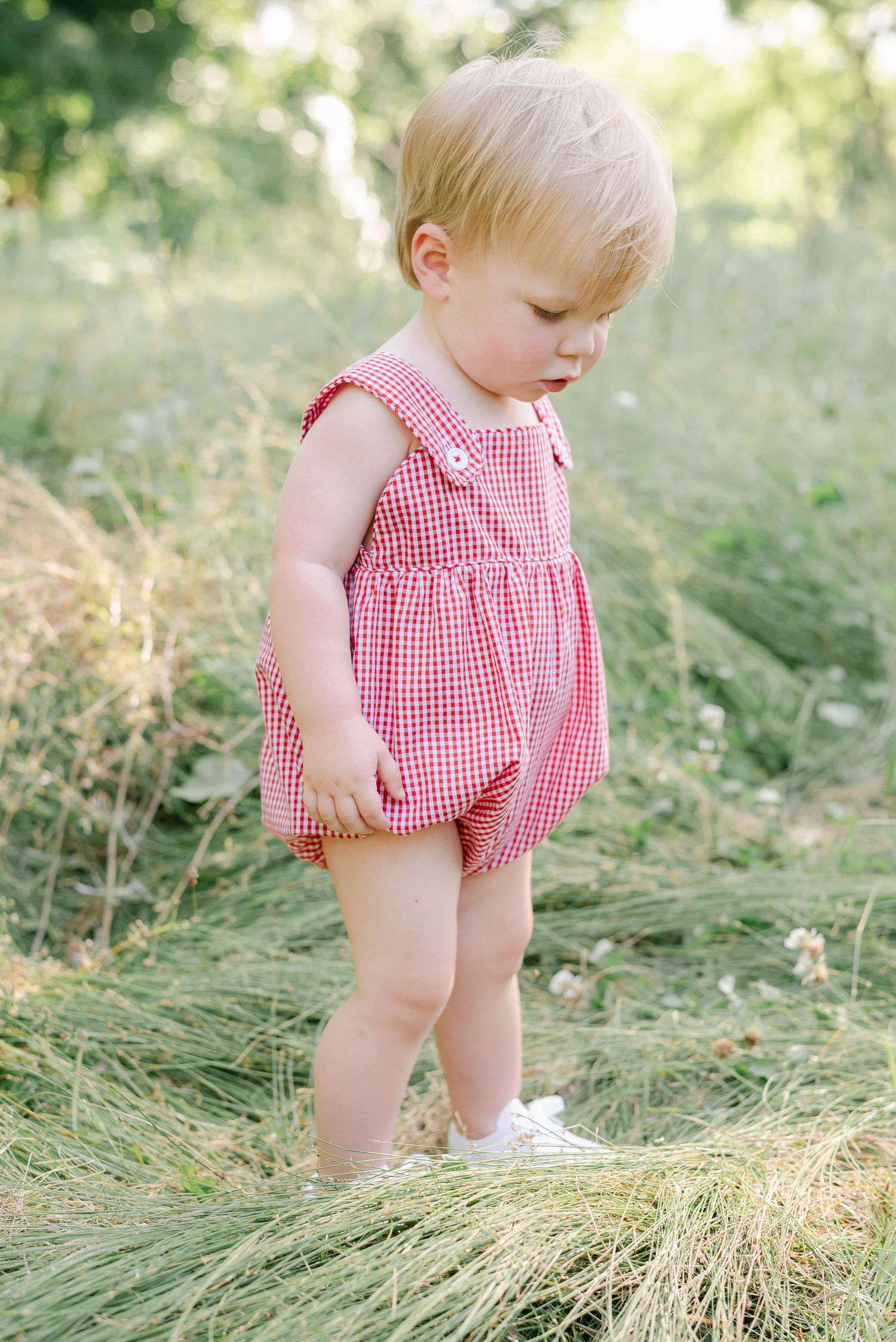 Baby in red gingham romper exploring outdoor grassy setting in summer.