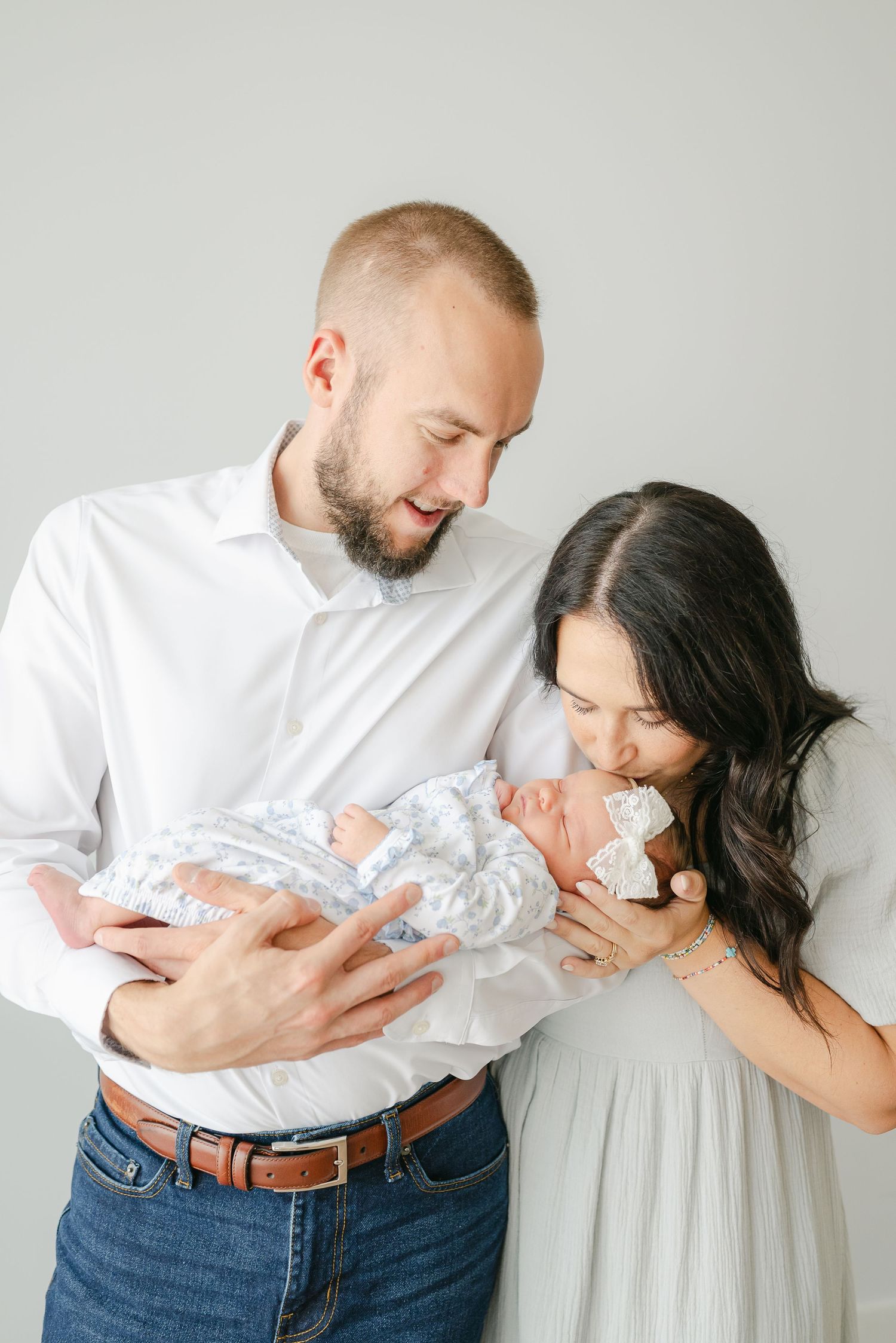 Intimate family portrait series with parents cradling newborn against grey backdrop.