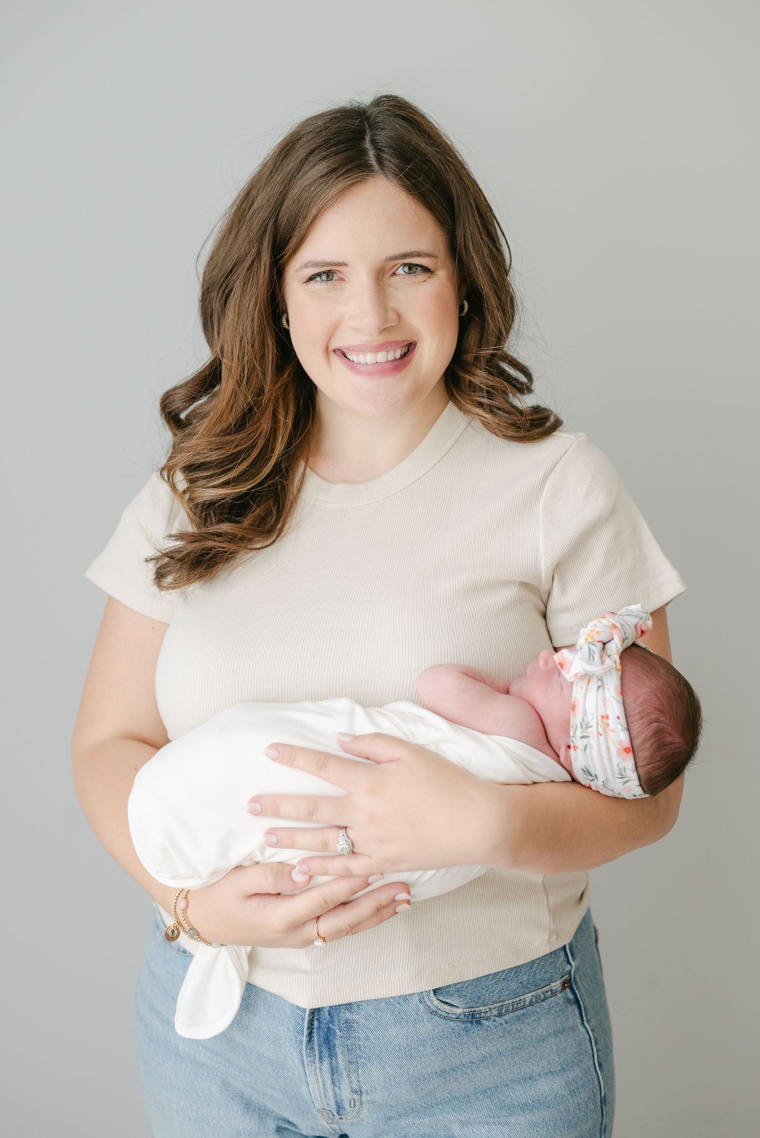 A new mother in a cream t-shirt and jeans cradles her newborn baby wrapped in white.
