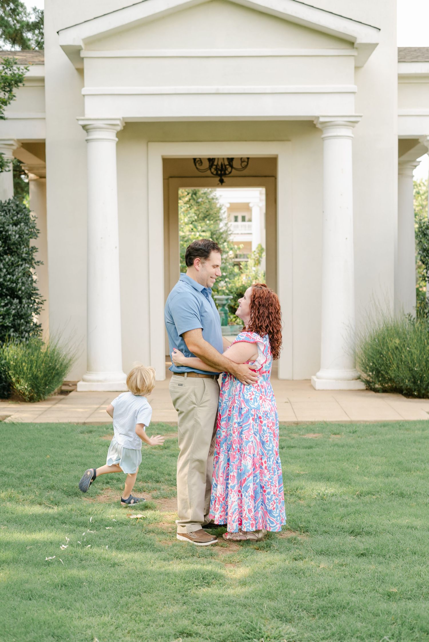 Family shares playful moments outside white columned building on green lawn.