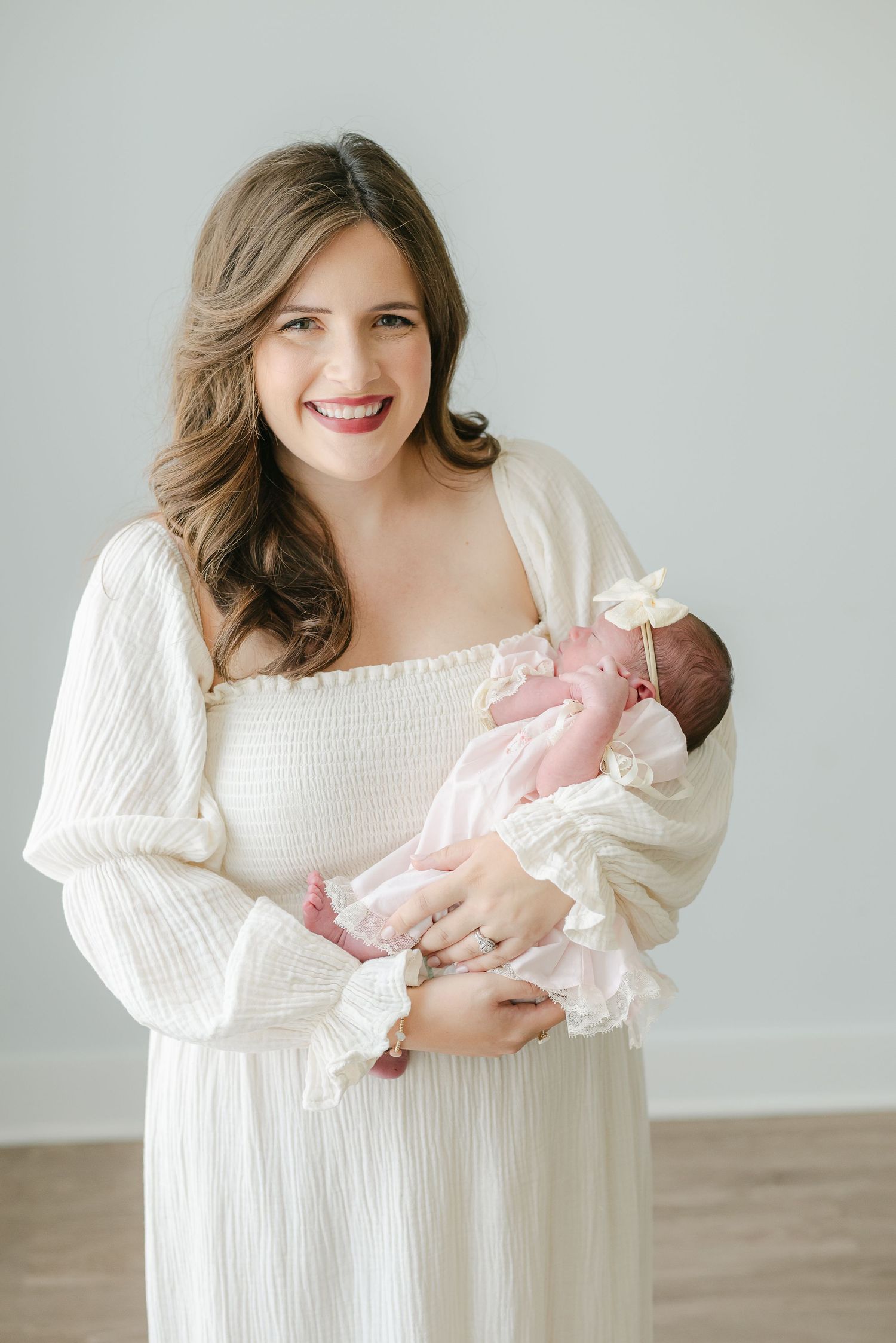 Woman in white dress holding newborn baby against minimal gray background.
