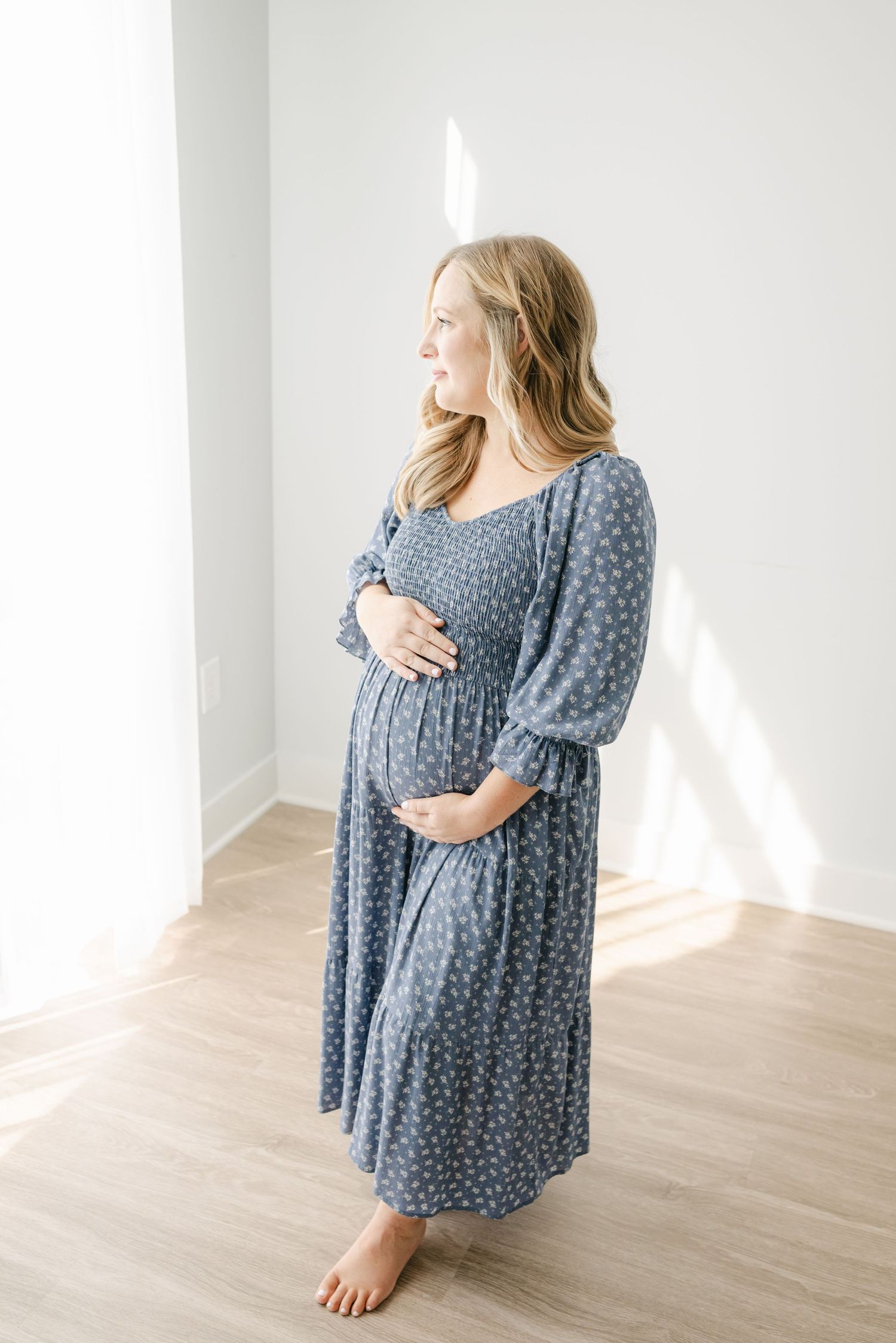Woman in blue floral maternity dress poses in sunlit studio setting.