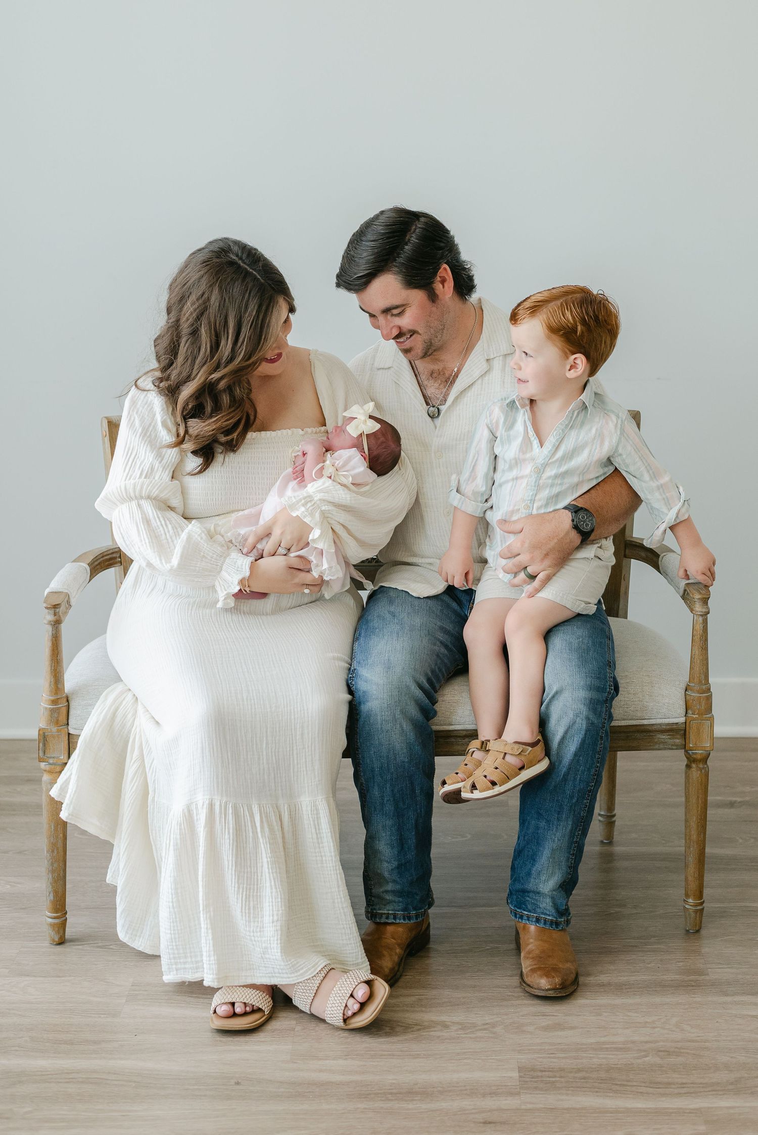 Family sitting together on wooden chairs in coordinated white outfits against light gray backdrop.