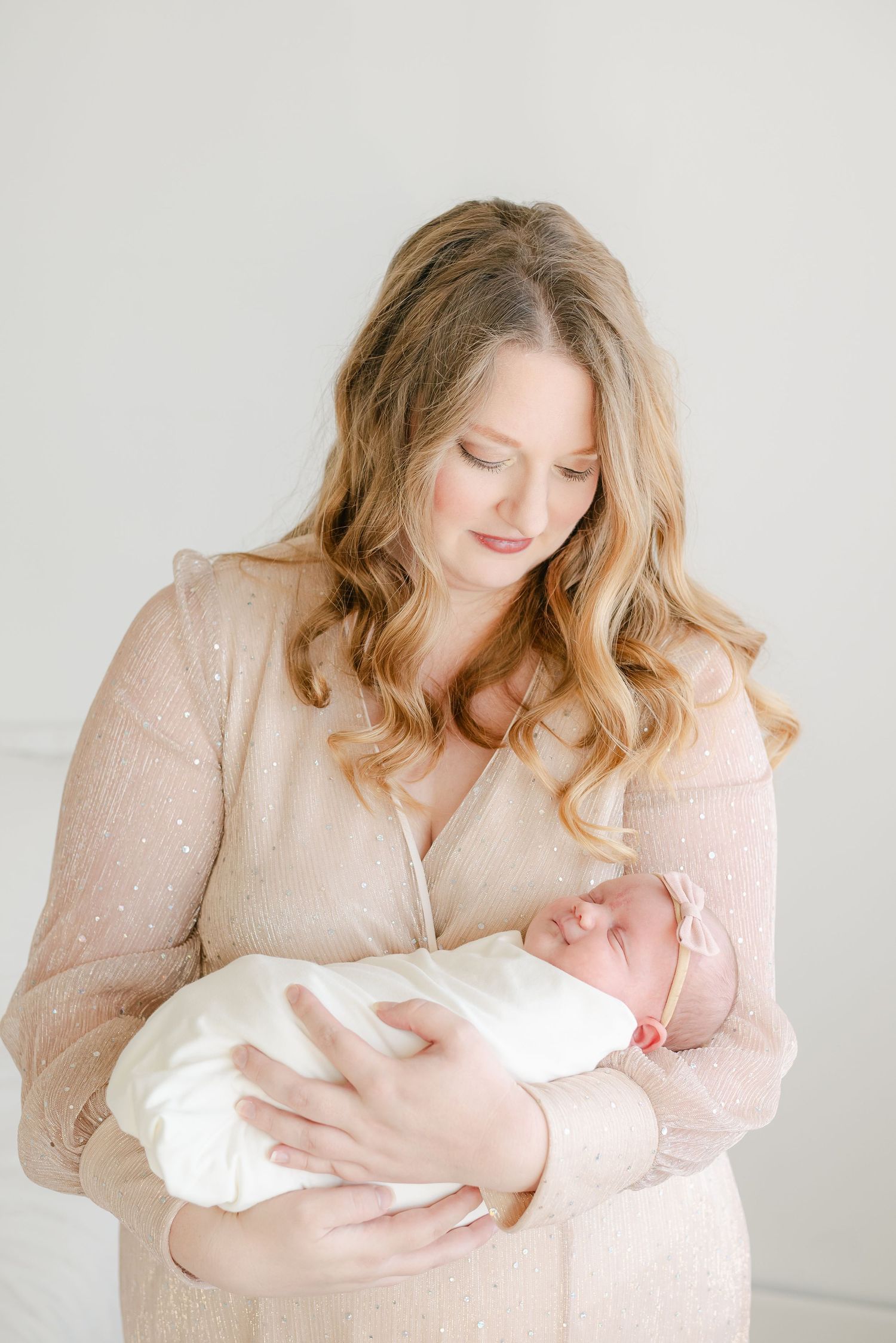 Mother in blush dress holding swaddled newborn against neutral background.