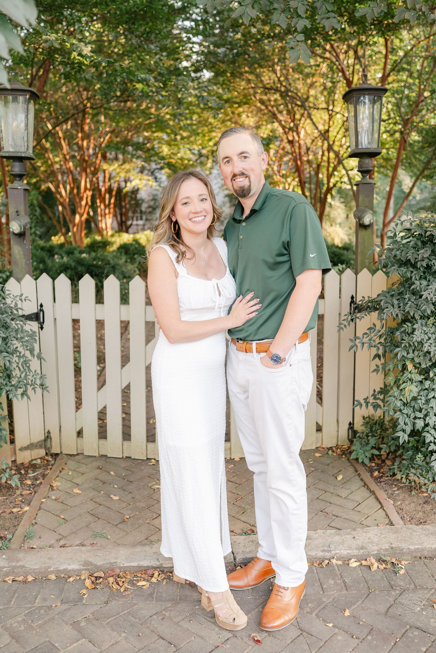 A couple in white outfits laughs together in front of a white picket fence.