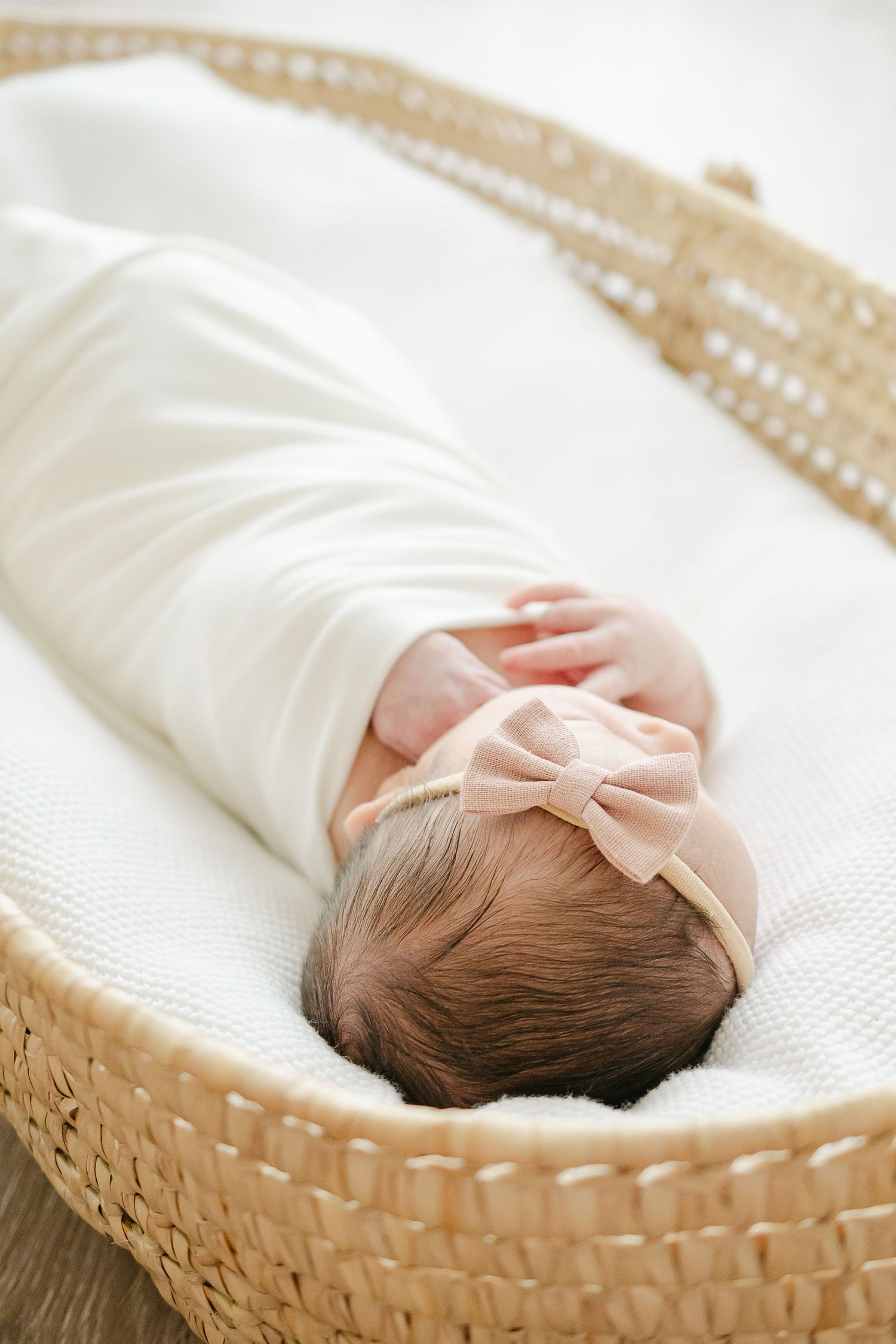 Newborn wrapped in white swaddle with bow headband lying in woven basket.