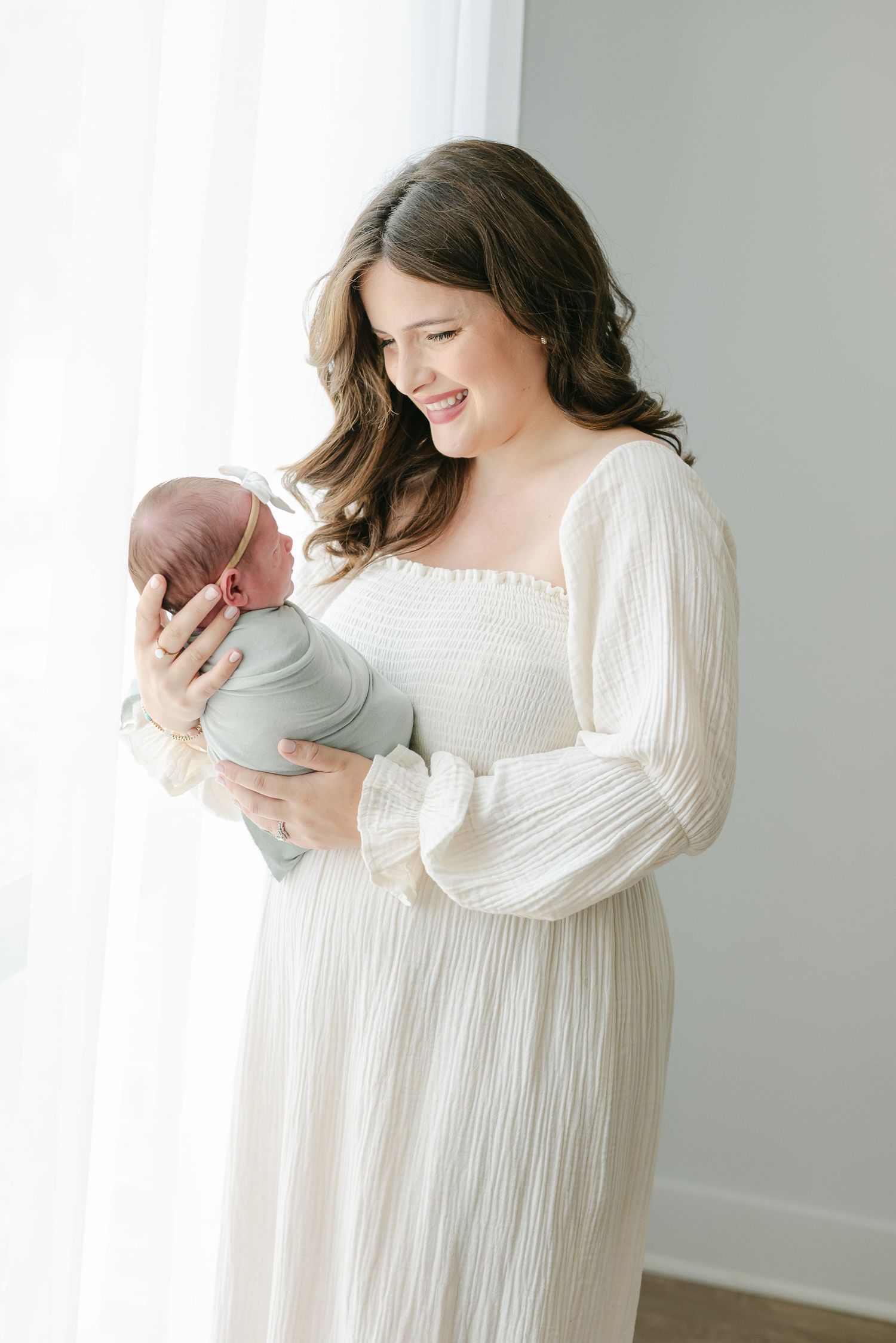 Woman in flowing white dress holding sleeping newborn in bright studio setting.