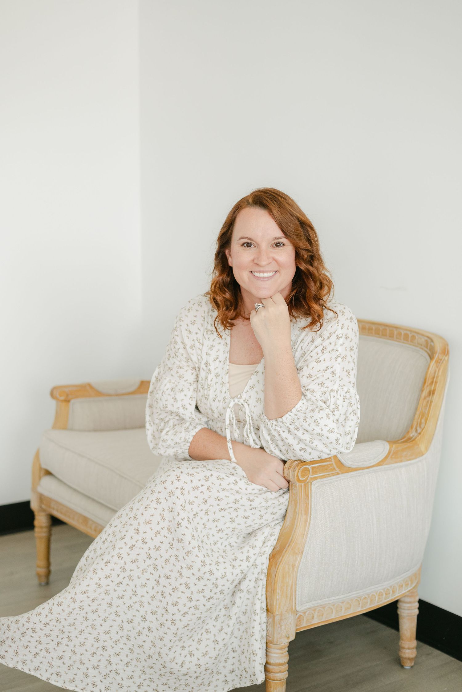 A person in a white floral dress poses casually on a beige chaise lounge against a light gray wall.