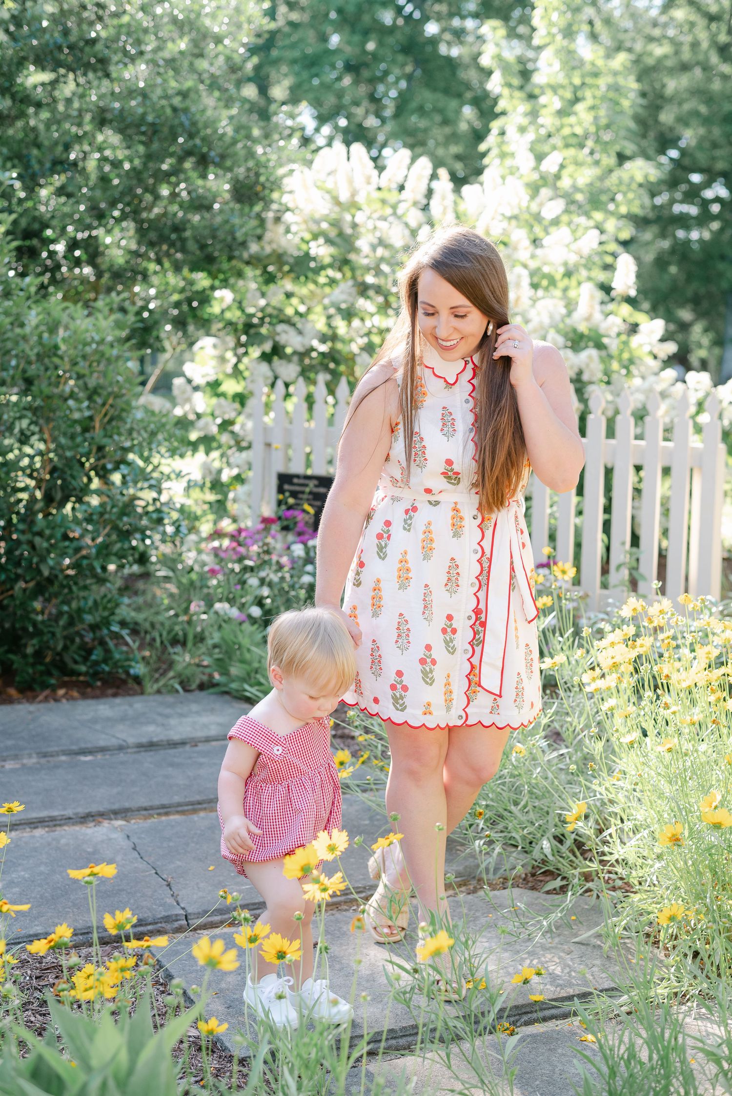 Parent and child exploring garden with yellow flowers along white picket fence.