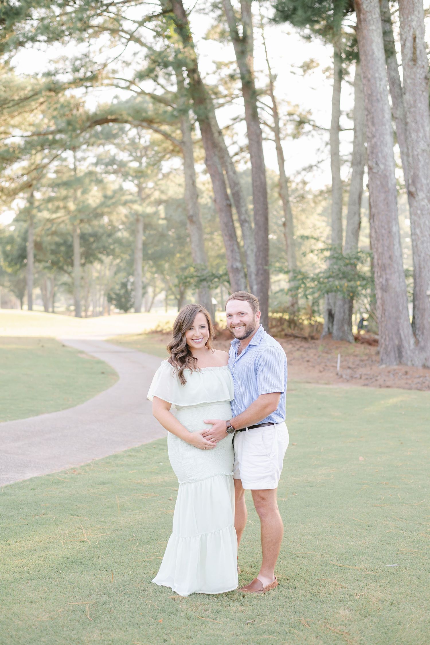 Romantic maternity photos of a couple in white outfits on a grassy path with pine trees.