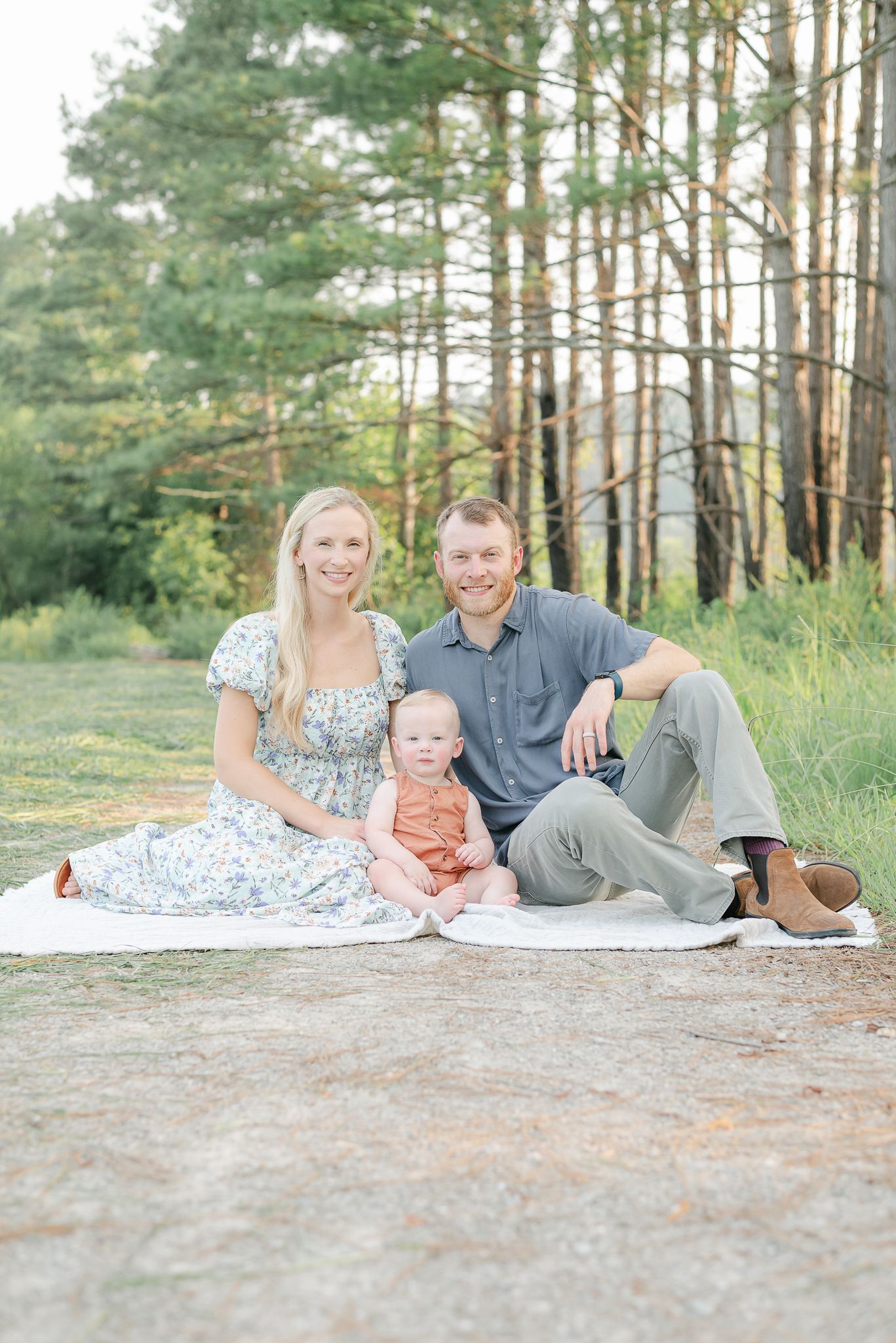 A family sits together on a white blanket in a serene pine forest setting during a summer photoshoot.