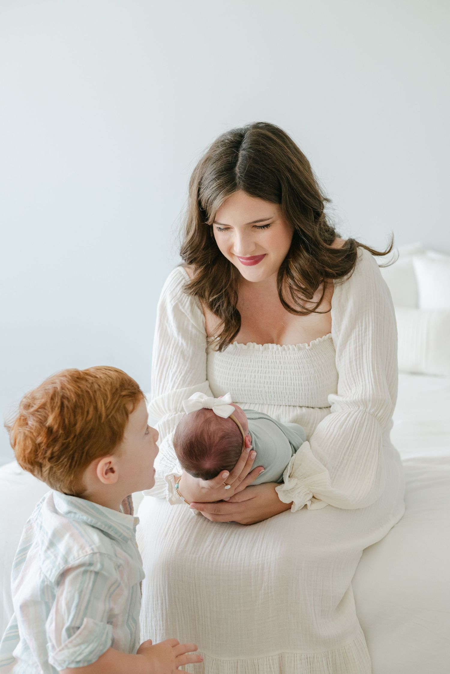 Mother in white dress sharing tender moment with children against light background.