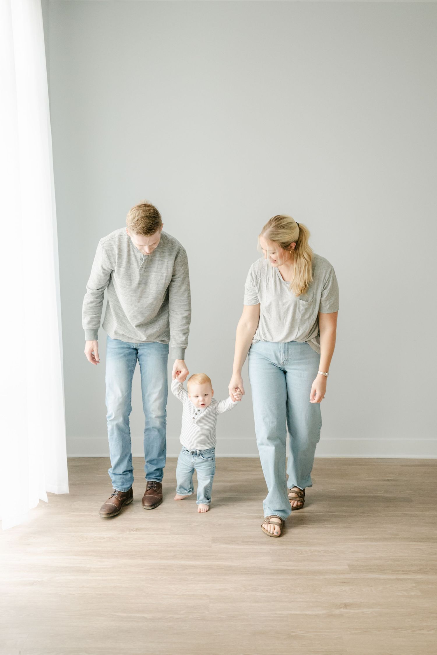 Family walking together holding hands in matching casual outfits on hardwood floor.