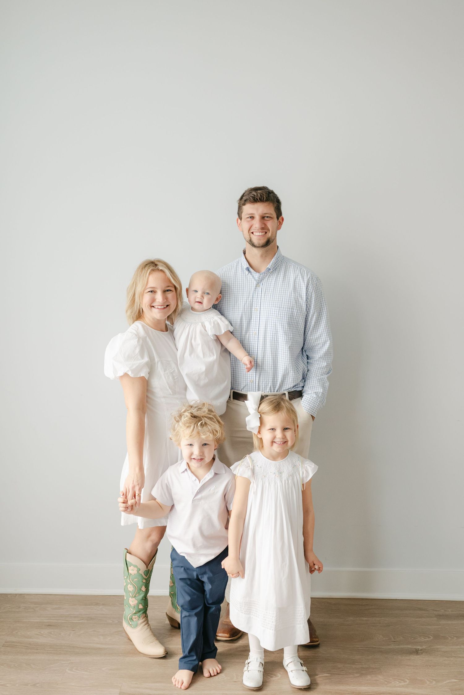 Family wearing coordinated white outfits poses against neutral wall for portrait session.