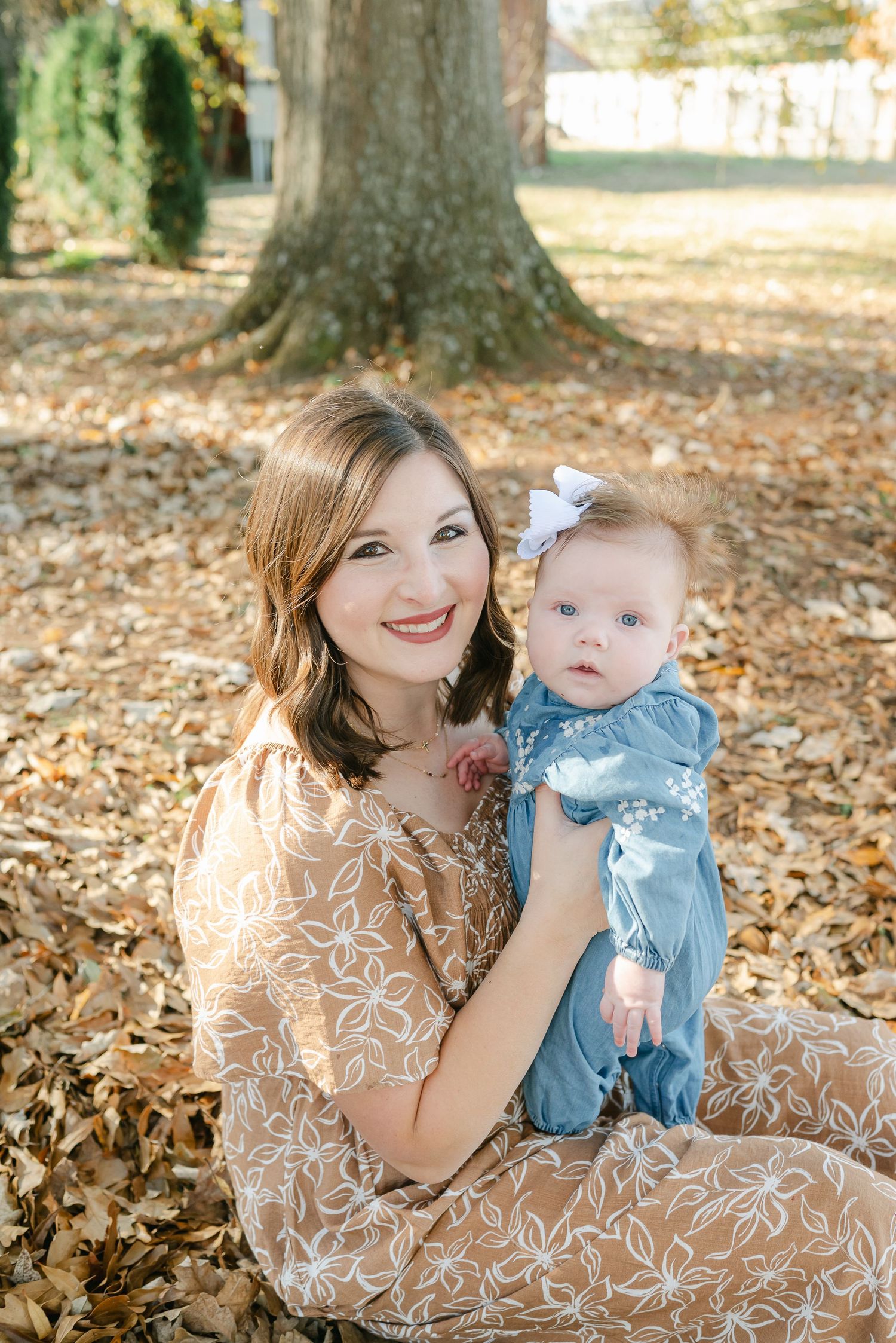 A parent and baby sit together on fallen autumn leaves in a park during a sunny outdoor photo session.