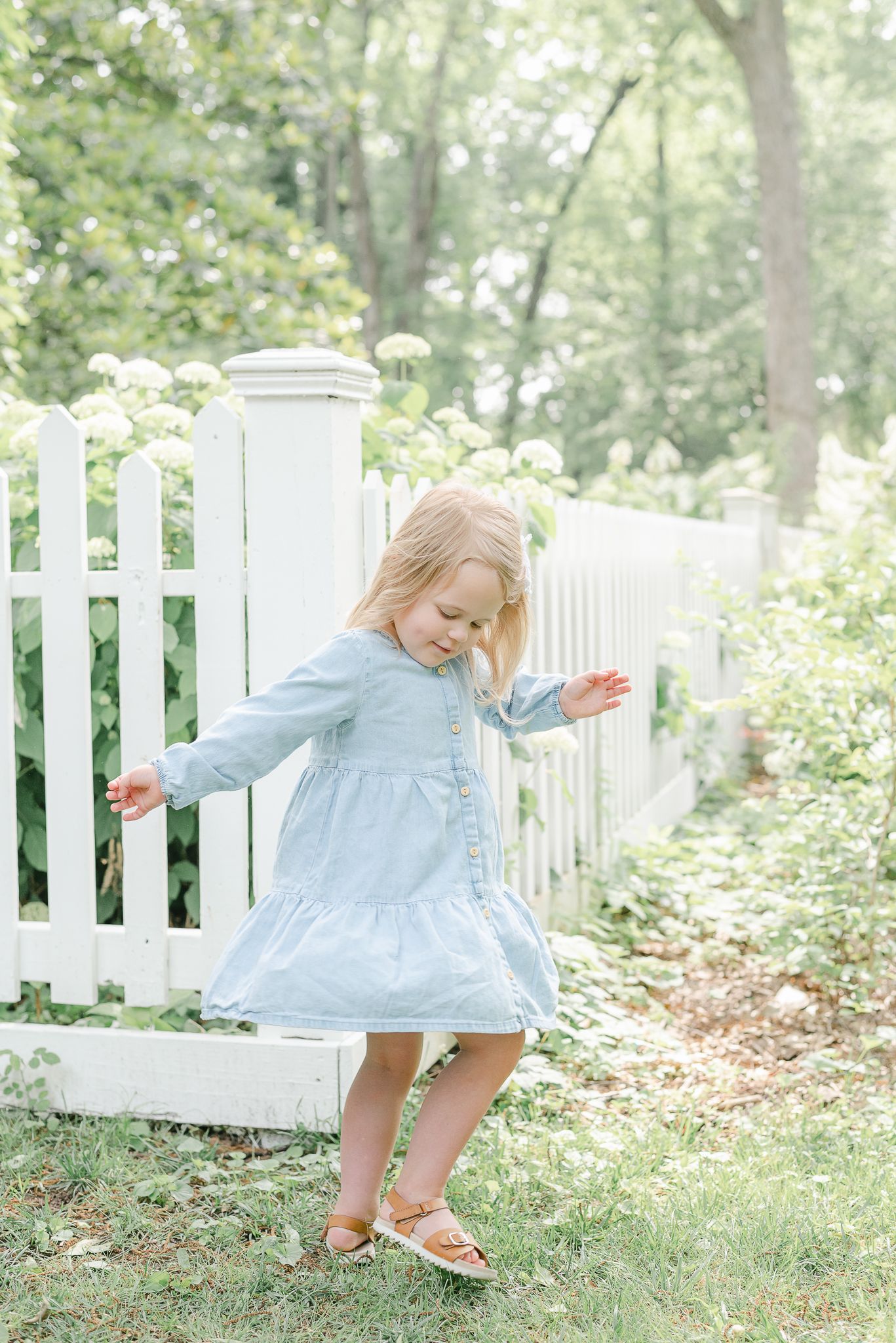 A young child in a light blue dress twirls playfully near a white picket fence in a garden setting.