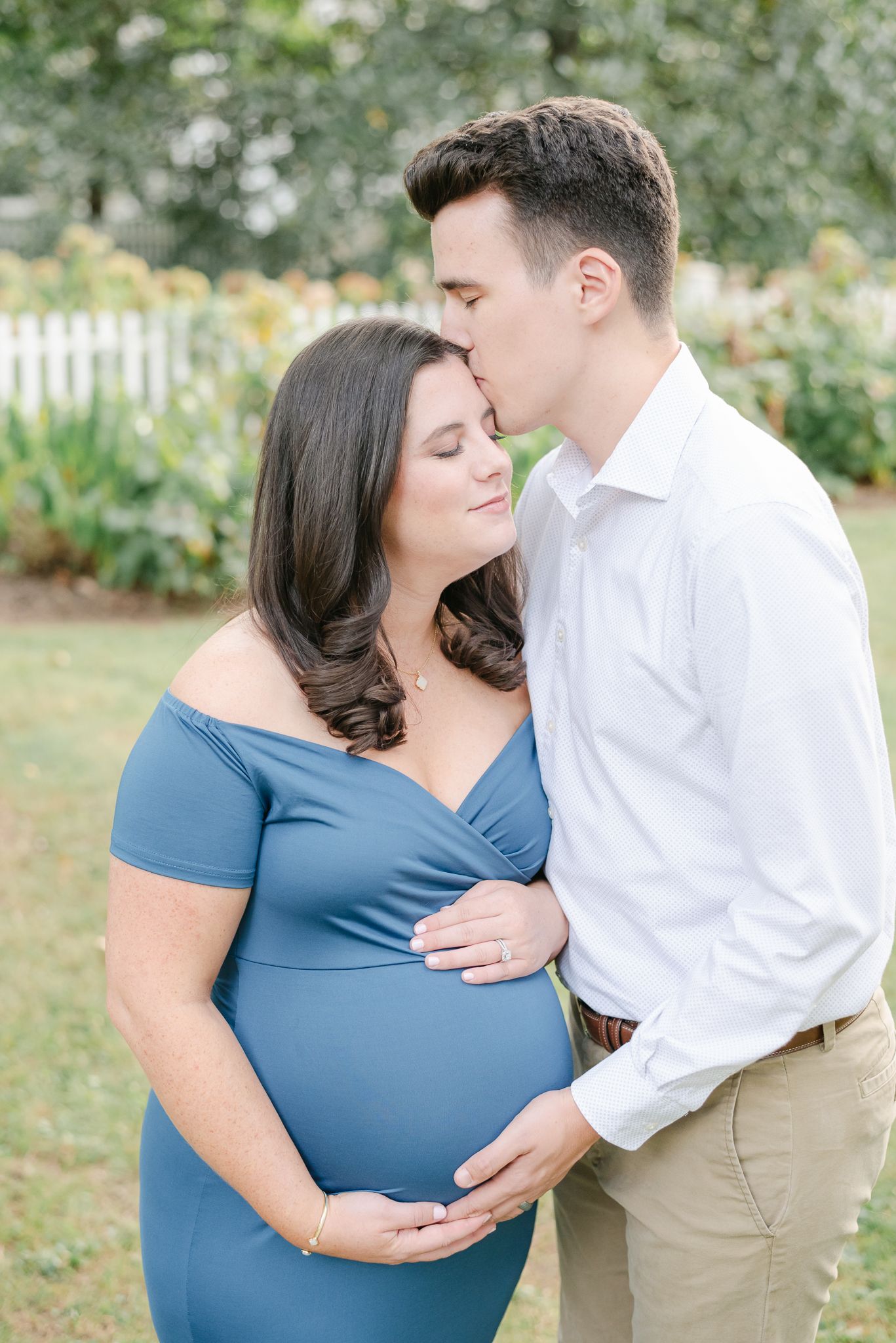 Romantic maternity portrait of couple in blue dress and white shirt against garden backdrop.