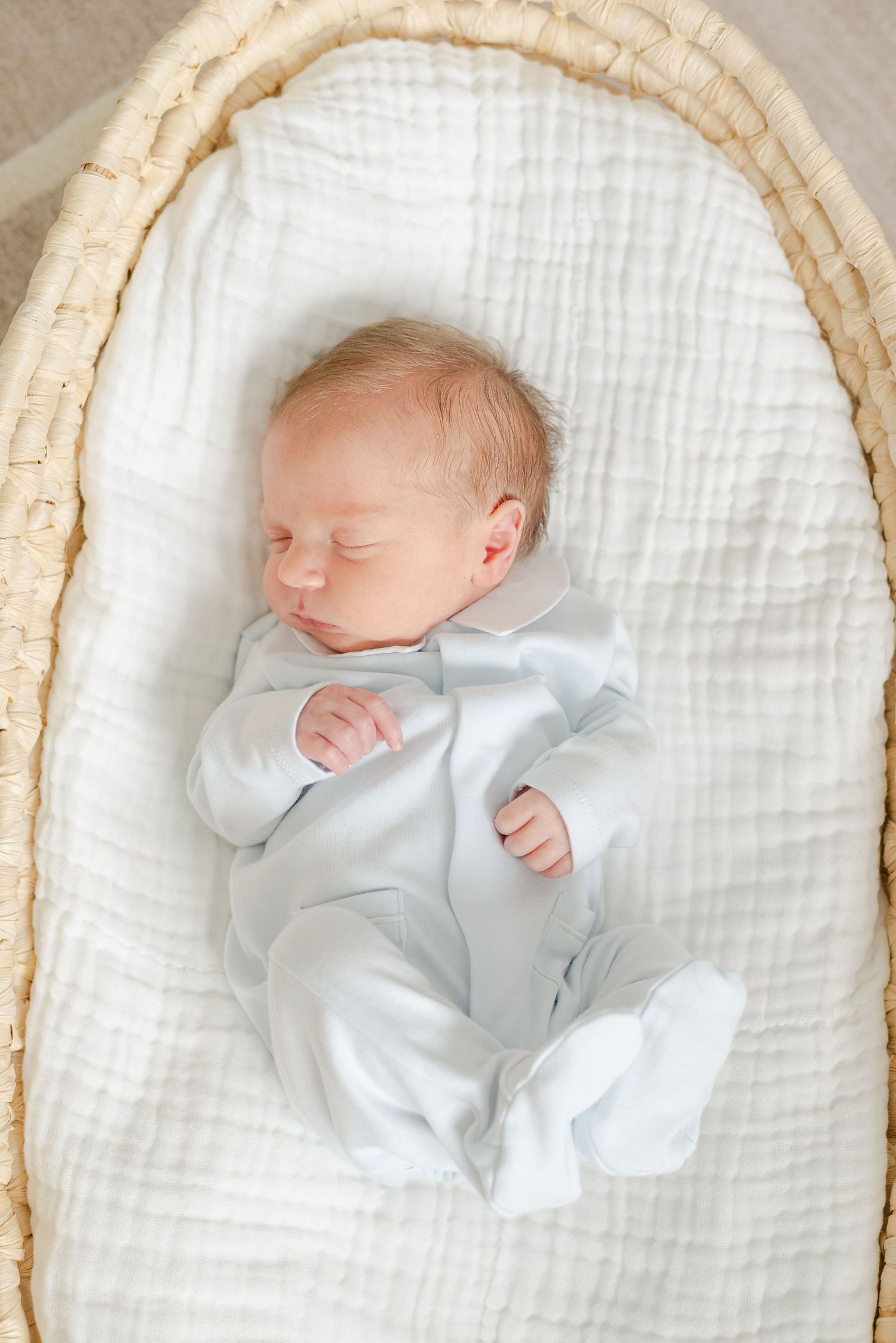 Sleeping newborn in white sleepwear resting in a cream-colored bassinet.