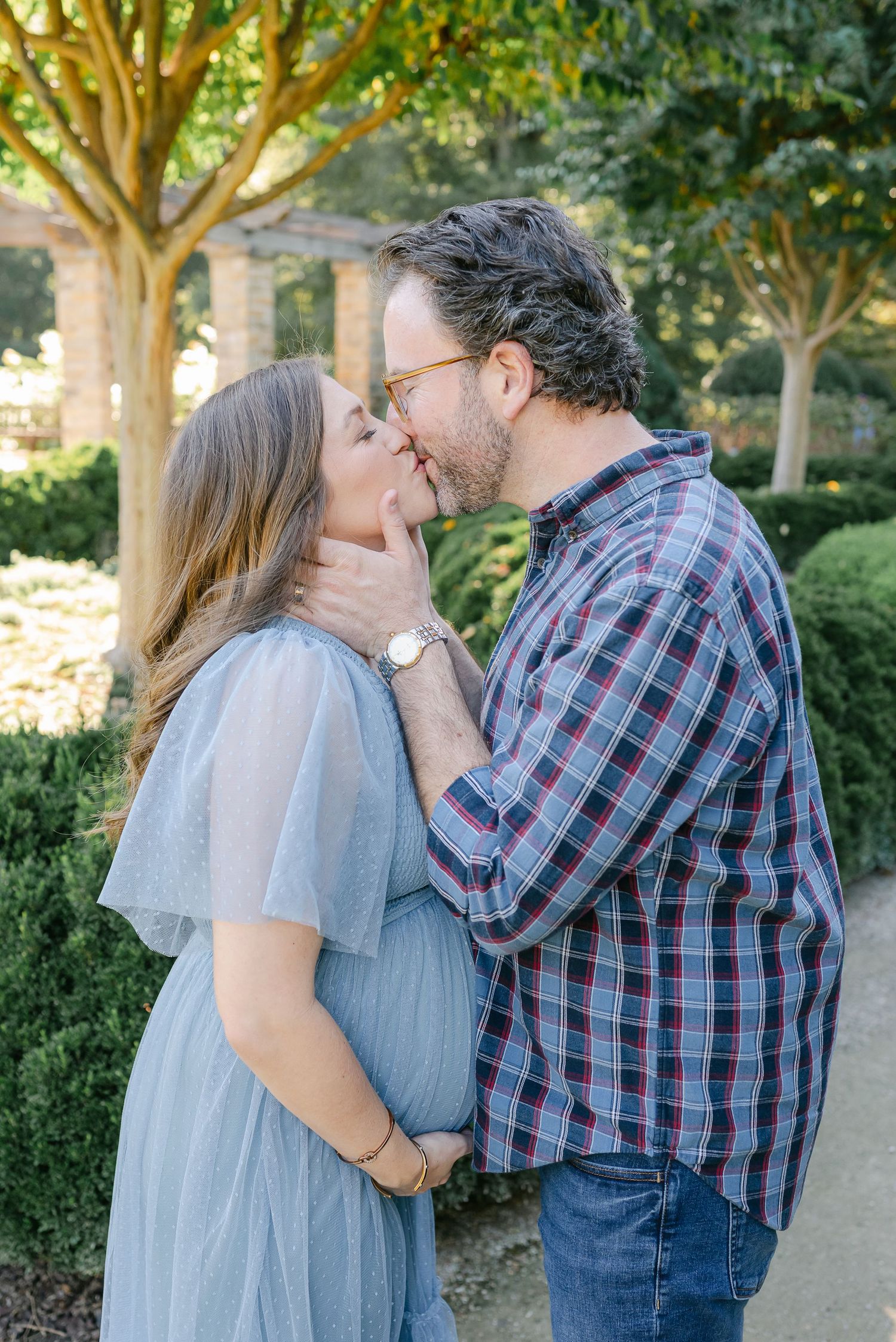 A couple shares an intimate kiss outdoors under trees in a garden setting.
