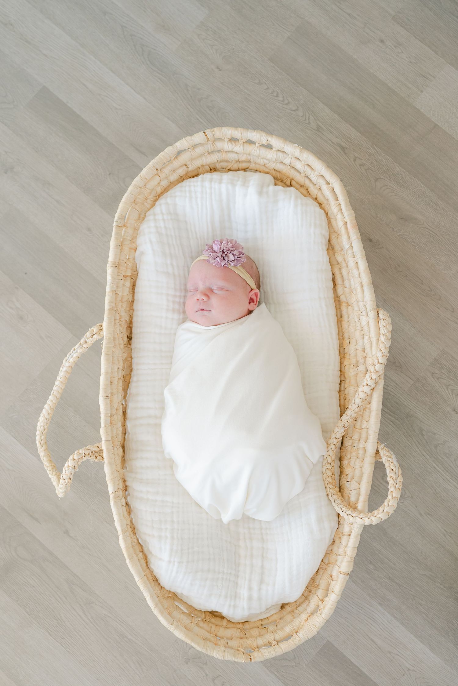 Newborn wrapped in white swaddle with pink headband sleeping in woven moses basket.