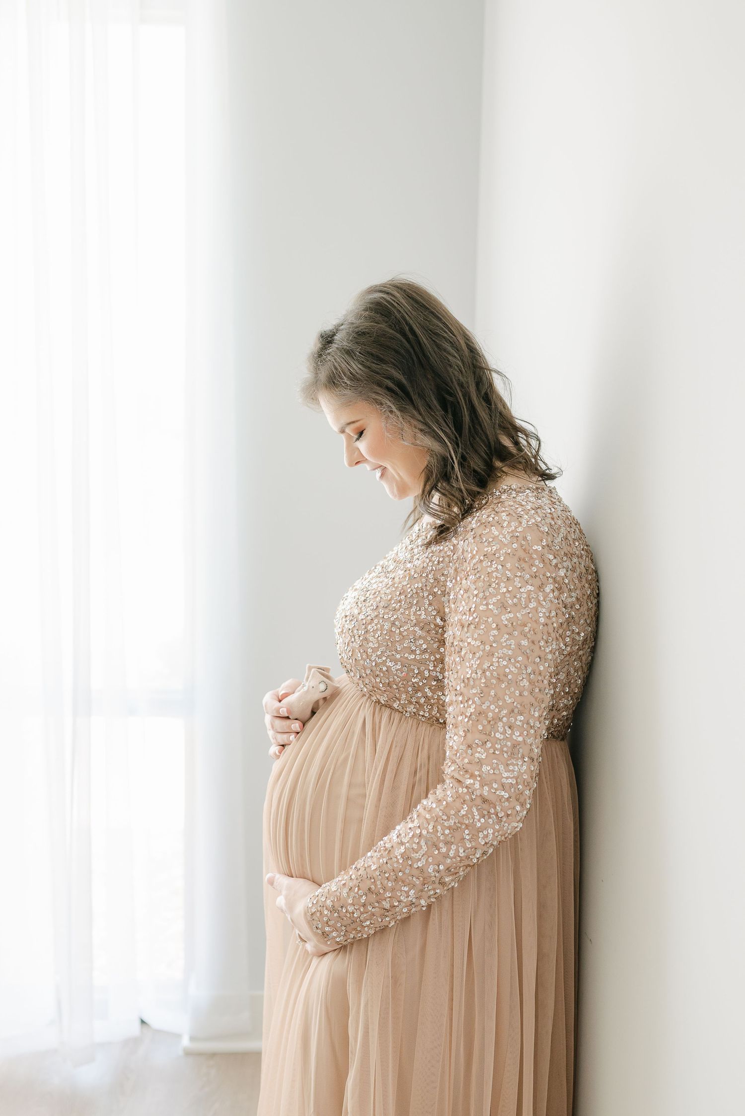 Expectant mother in a champagne-colored sequined maternity gown poses against a white wall.