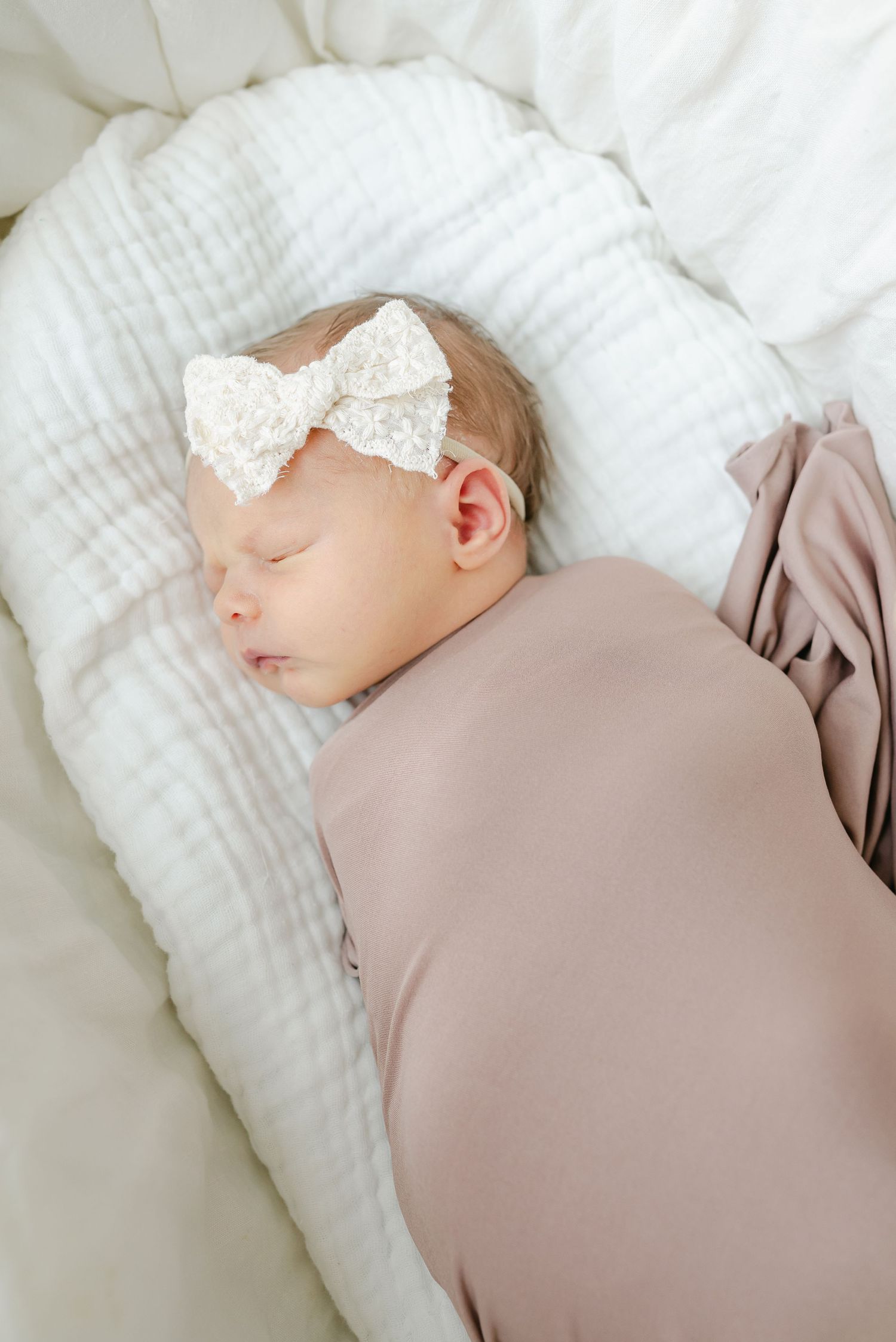 Sleeping newborn with lace bow headband wrapped in mauve blanket on white bedding.
