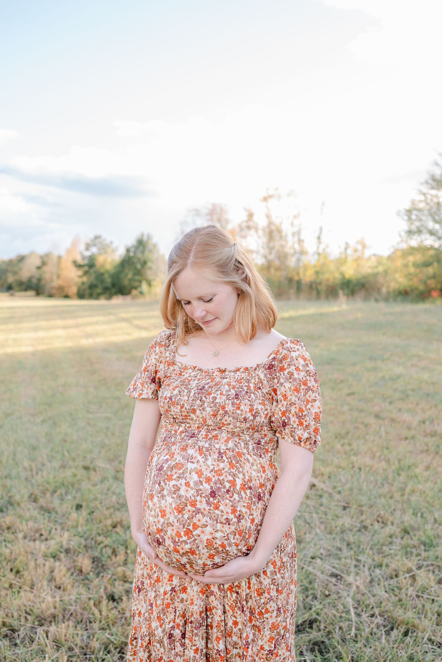 A pregnant woman in a floral orange dress stands in a sunlit field during golden hour.