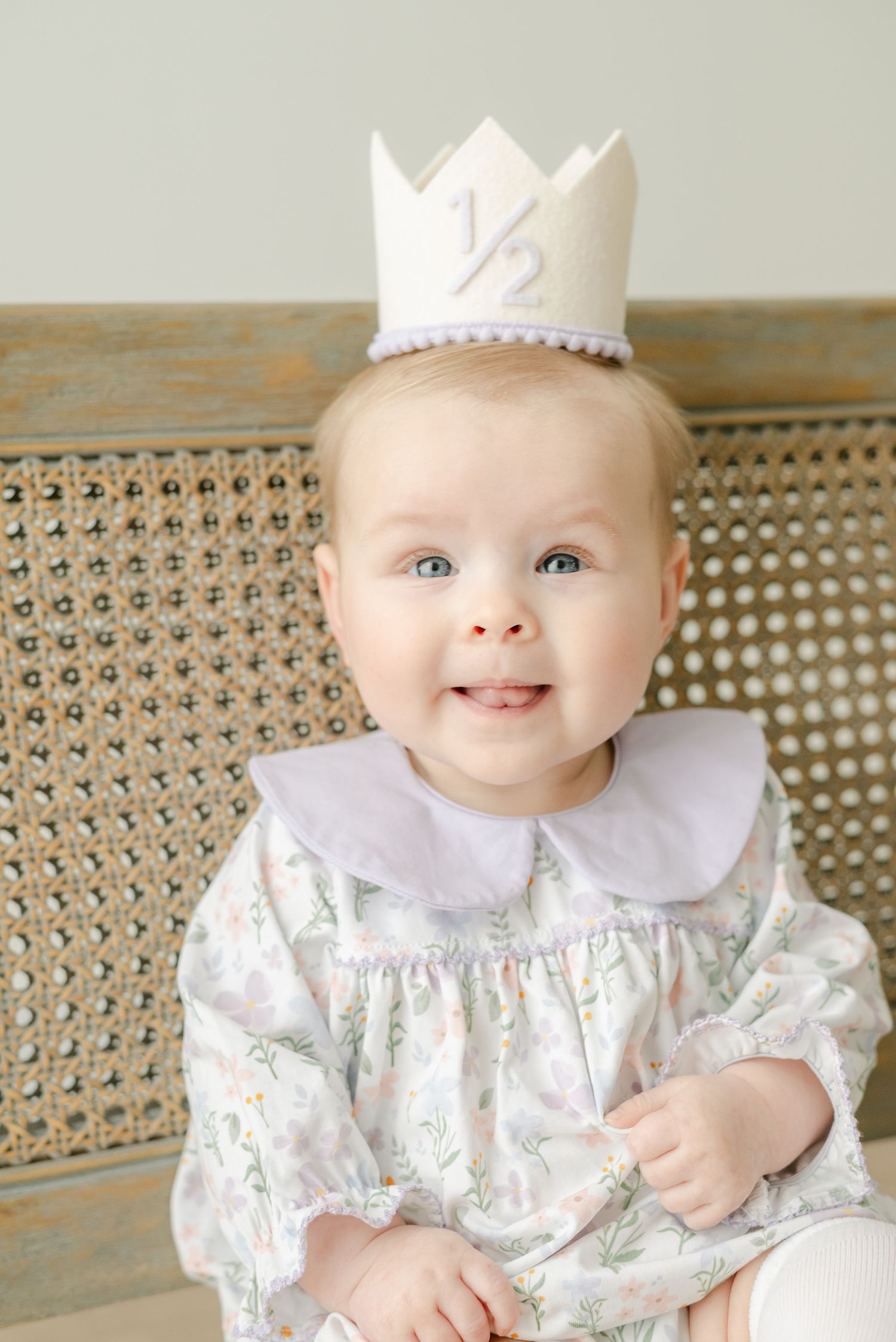 Baby wearing white dress and crown sitting on wicker chair in studio setting.