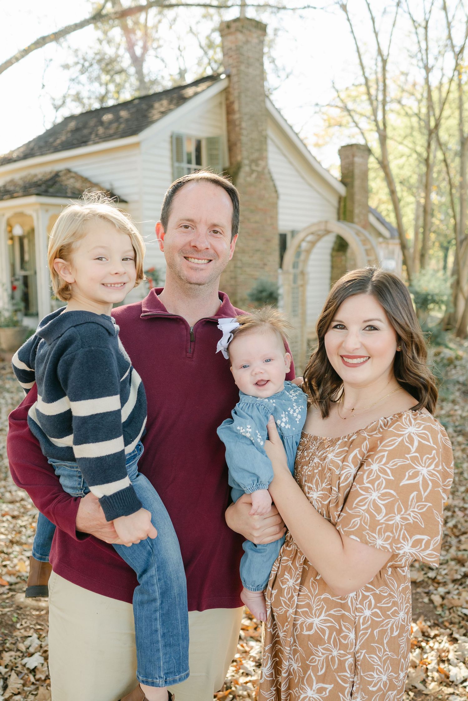 A family in coordinated outfits poses together in a wooded setting with autumn leaves on the ground.