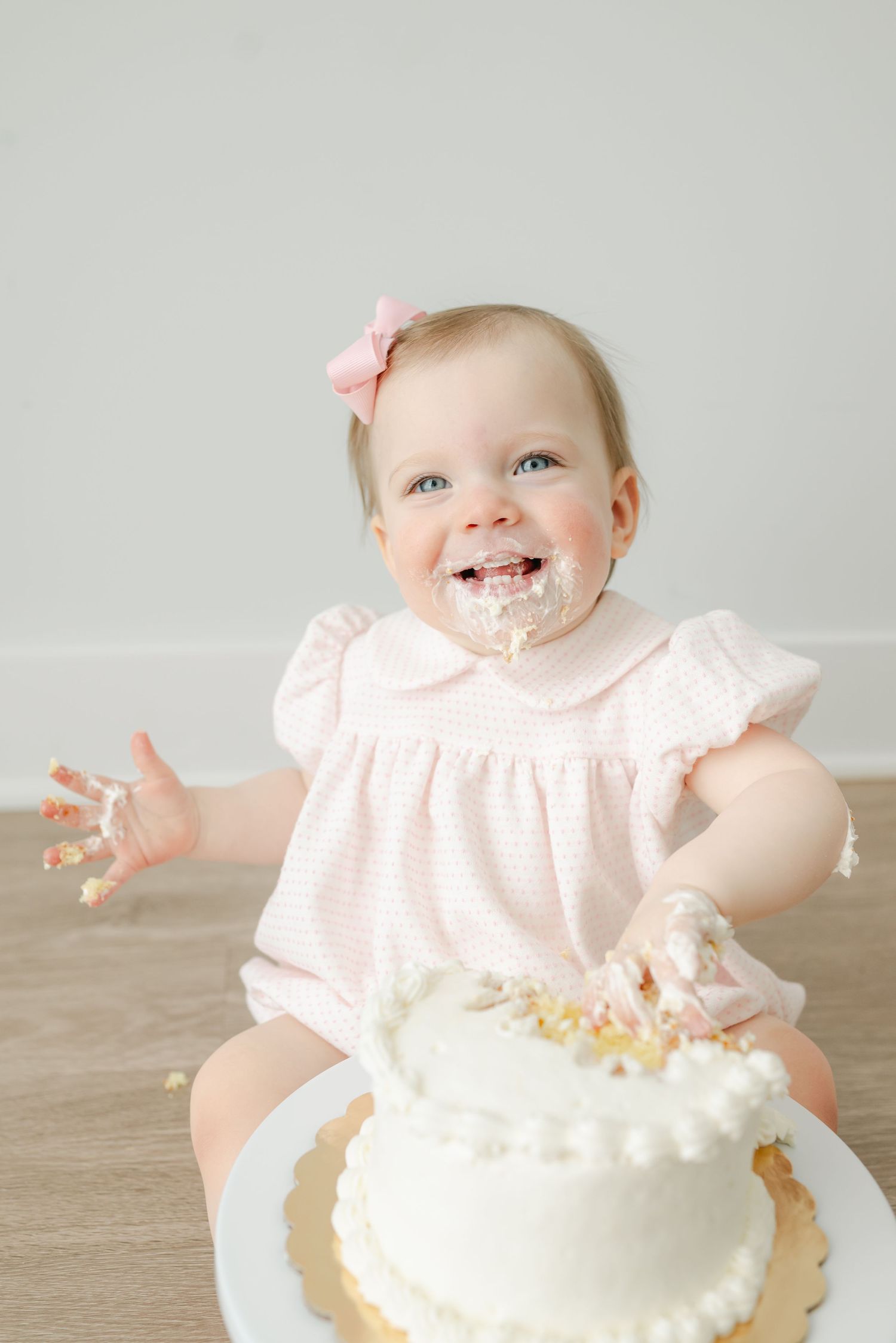 Baby in pink dress laughing with cake frosting on face during cake smash photo session.