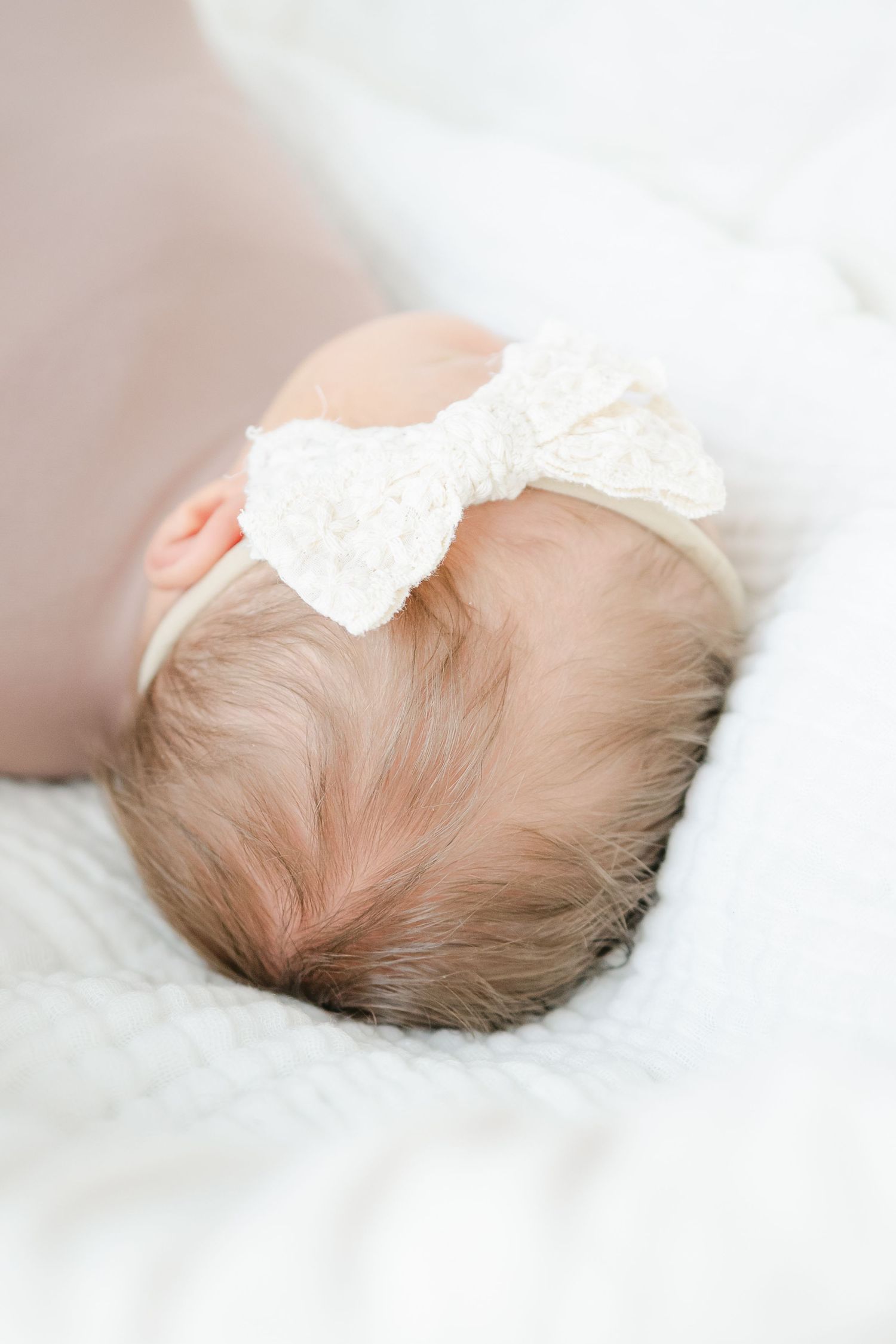 Close-up profile of sleeping newborn with white lace bow on soft white fabric.