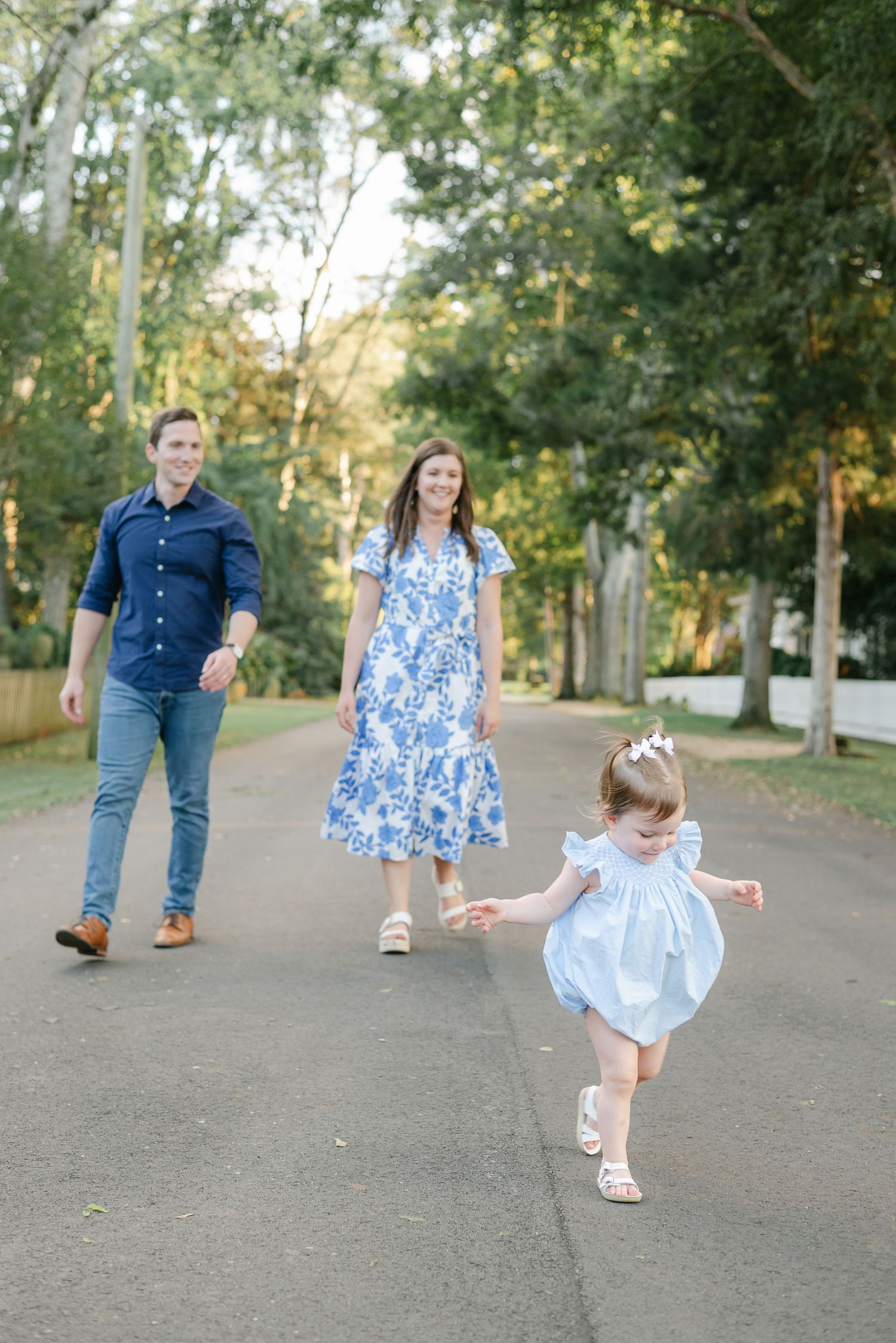 A family walks together on a tree-lined path as a young child in a light blue dress skips ahead playfully.