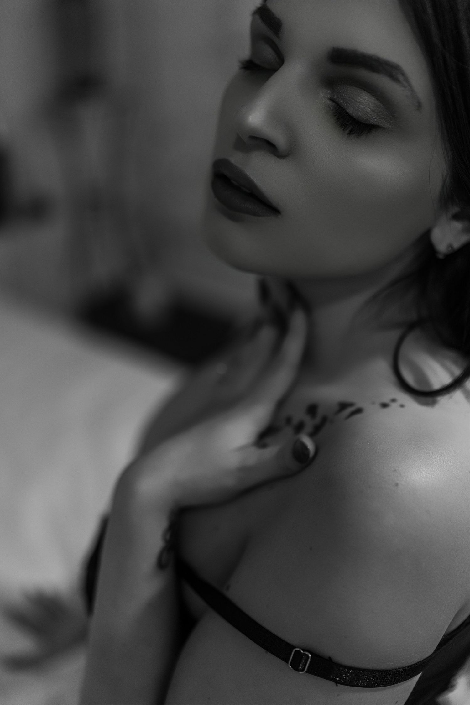 A black and white artistic portrait showing sultry makeup, closed eyes, dark lipstick and visible shoulder tattoo details.