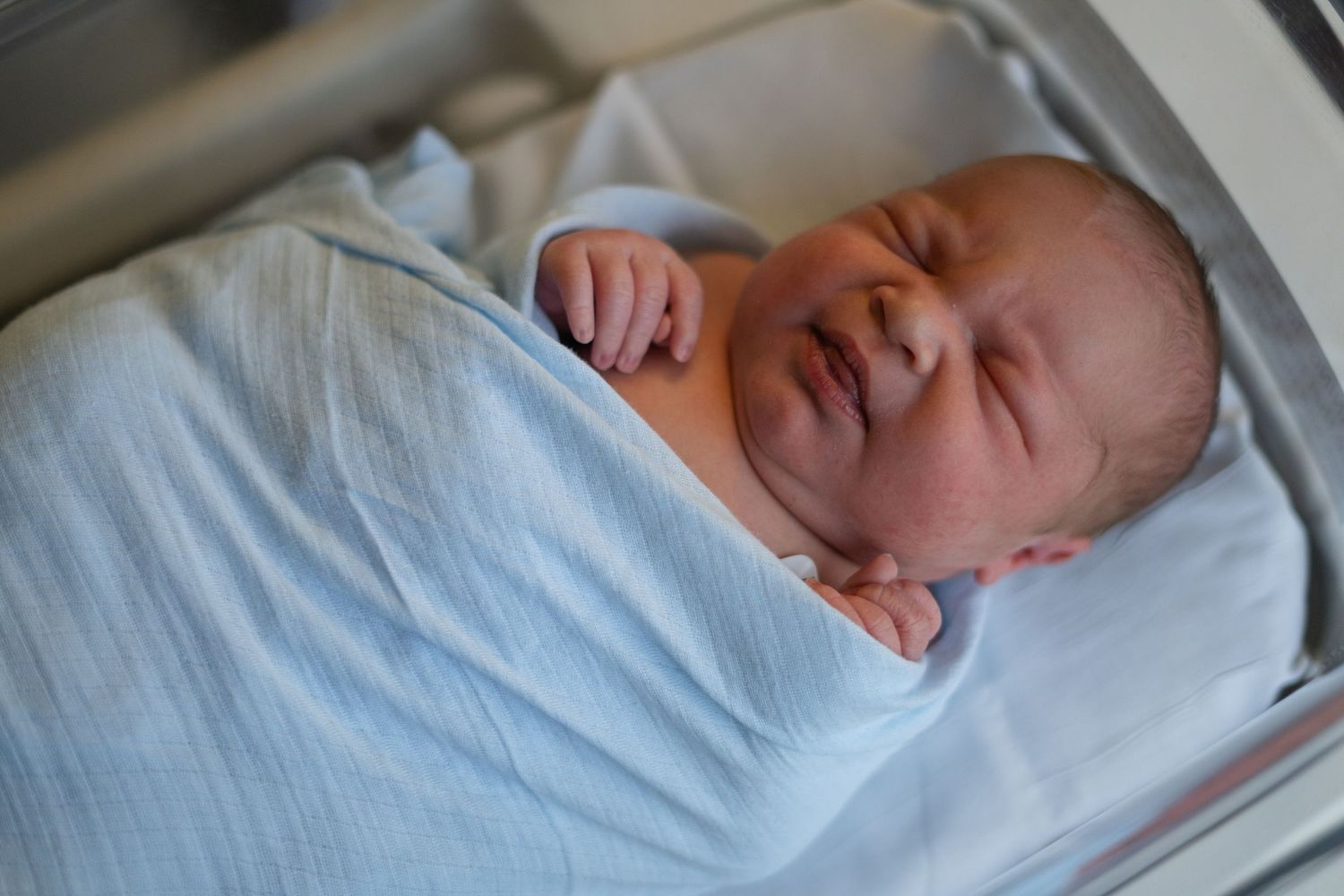 Sleeping newborn baby wrapped in white blanket lying on hospital bed while medical exam is performed.