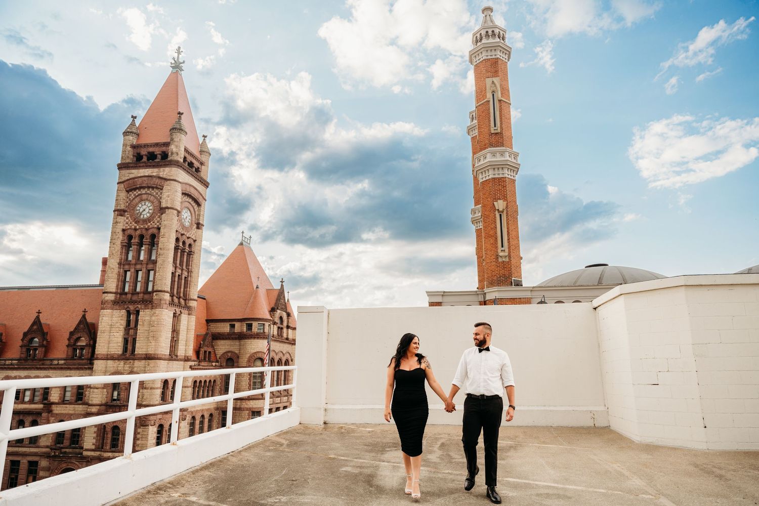 A couple walks along a white terrace overlooking a historic church spire and city skyline under a cloudy blue sky.