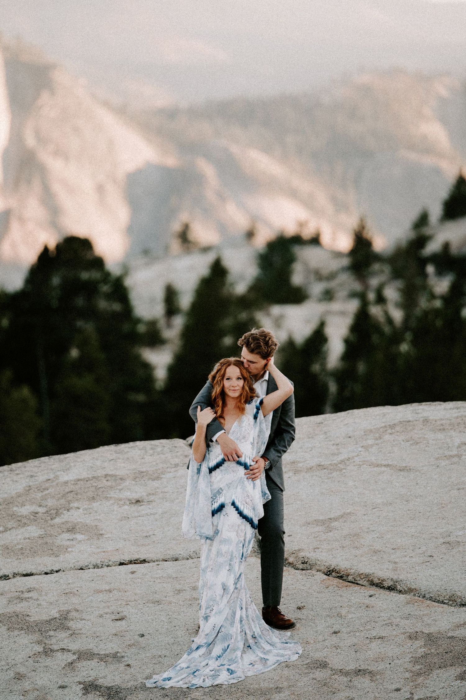 A romantic couple embraces on a snowy mountain slope at sunset with evergreen trees in the background.