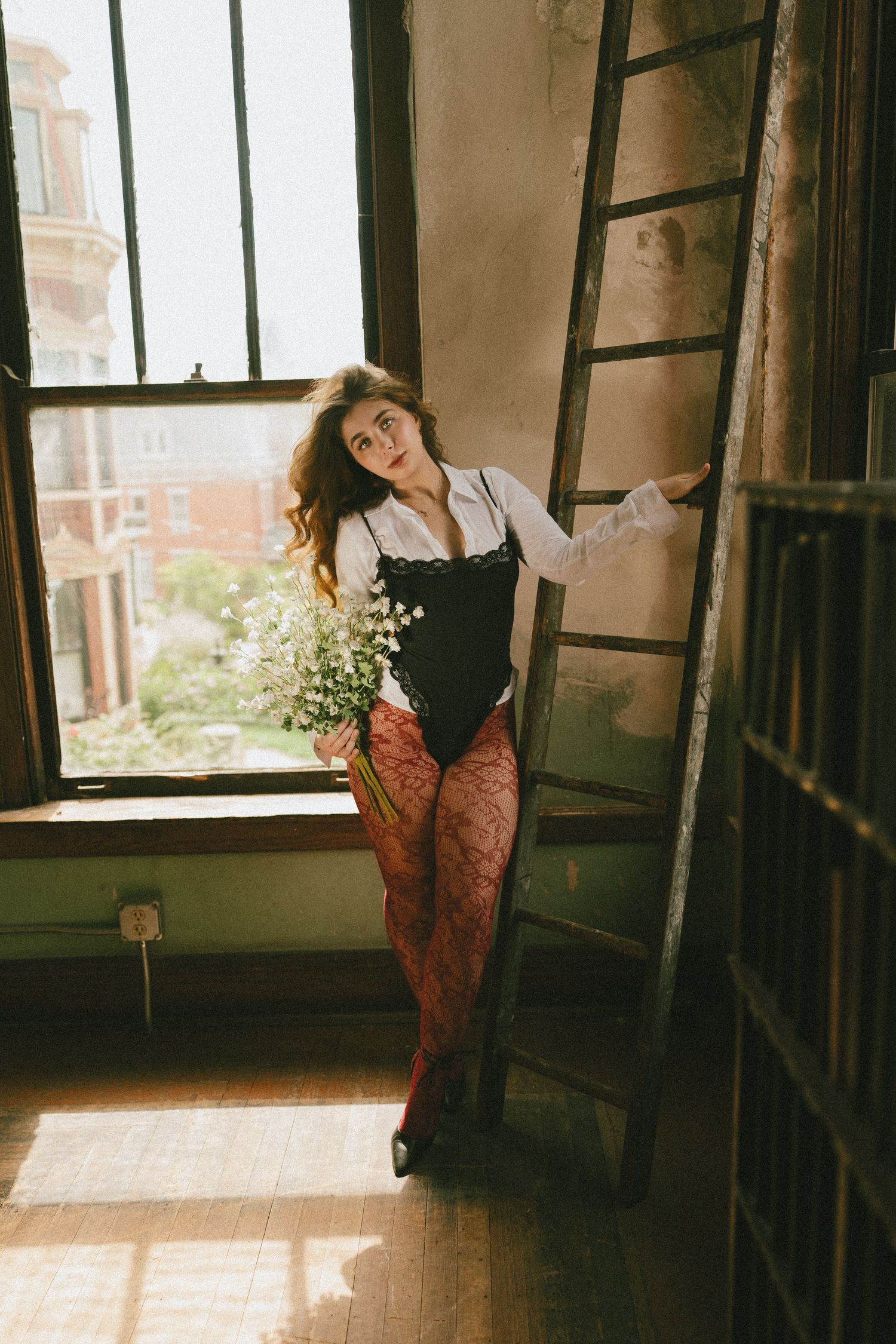 Woman in black corset and red lace tights holds white flowers by a sunlit window near a wooden ladder.