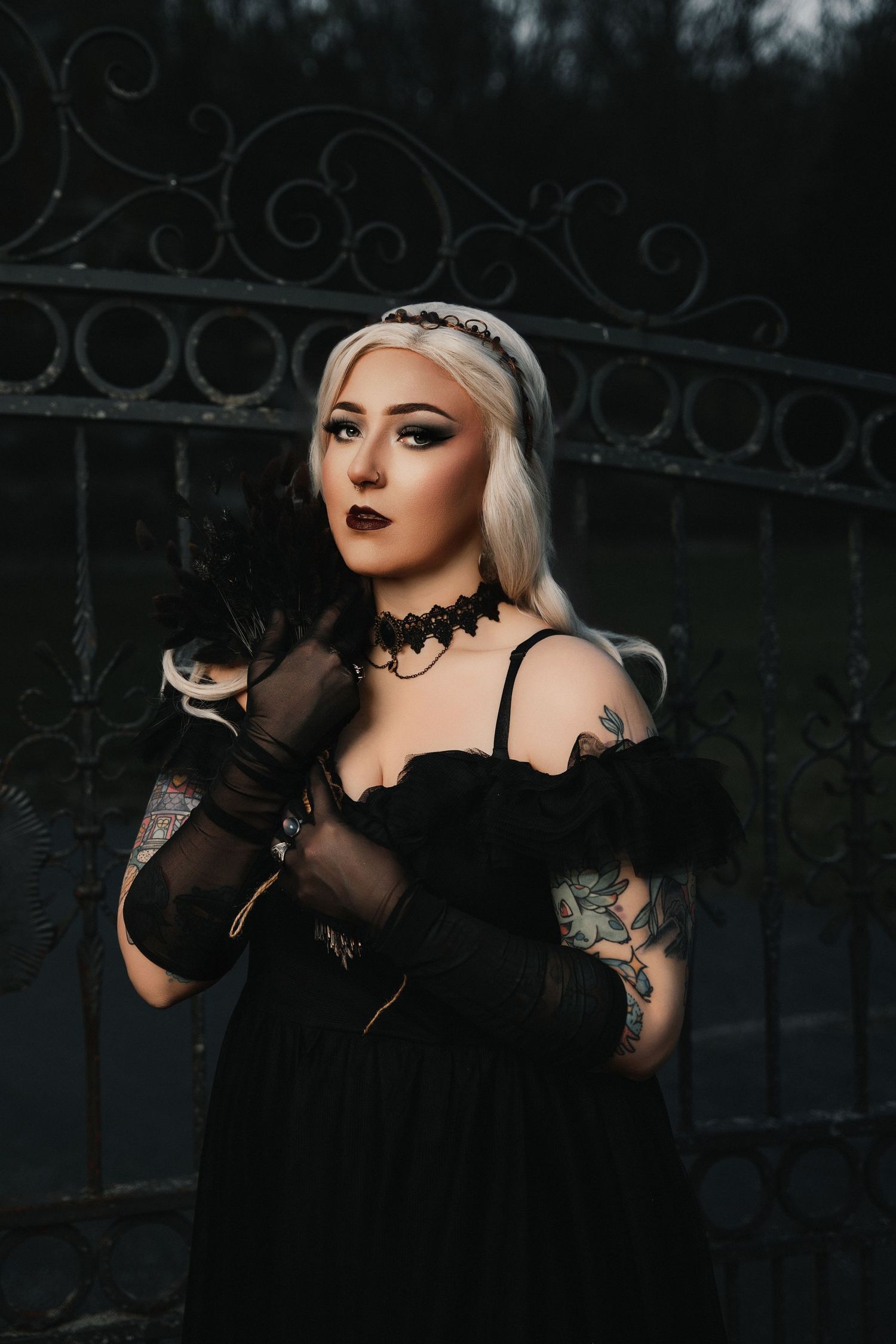 Gothic style portrait with black dress, lace choker, sheer gloves and tattoos against ornate metal gate at dusk.