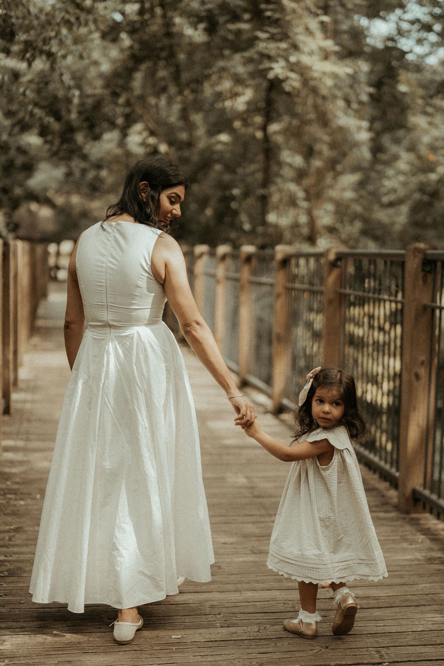 A mother and daughter in matching white dresses hold hands while walking on a wooden bridge in a lush park.