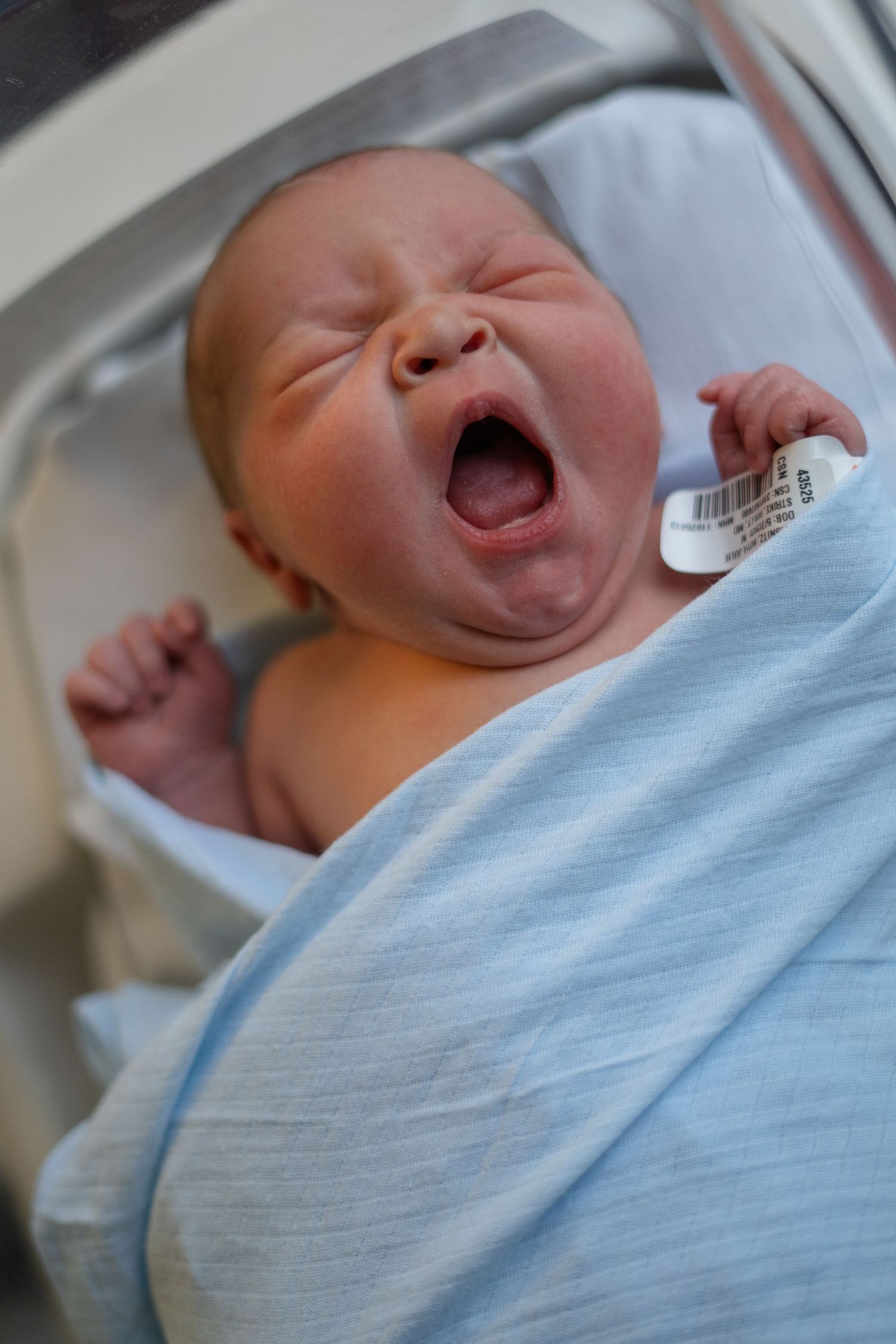Newborn baby yawning while wrapped in light blue hospital blanket.