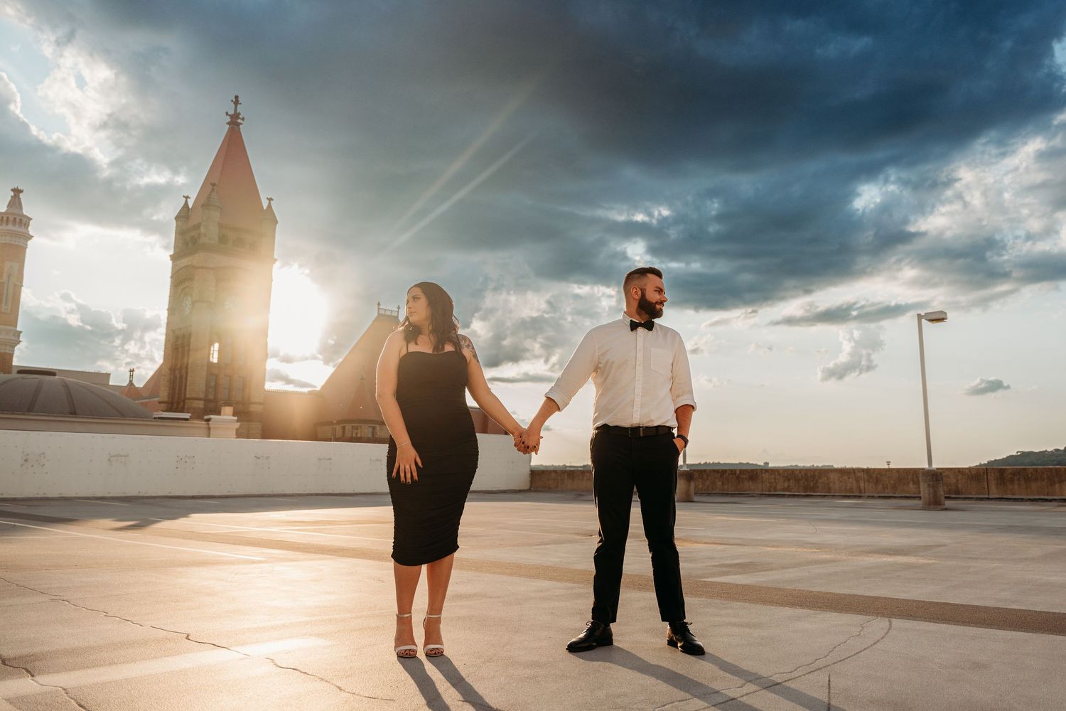 A couple holds hands on a rooftop terrace at sunset with a church tower silhouetted in the background.