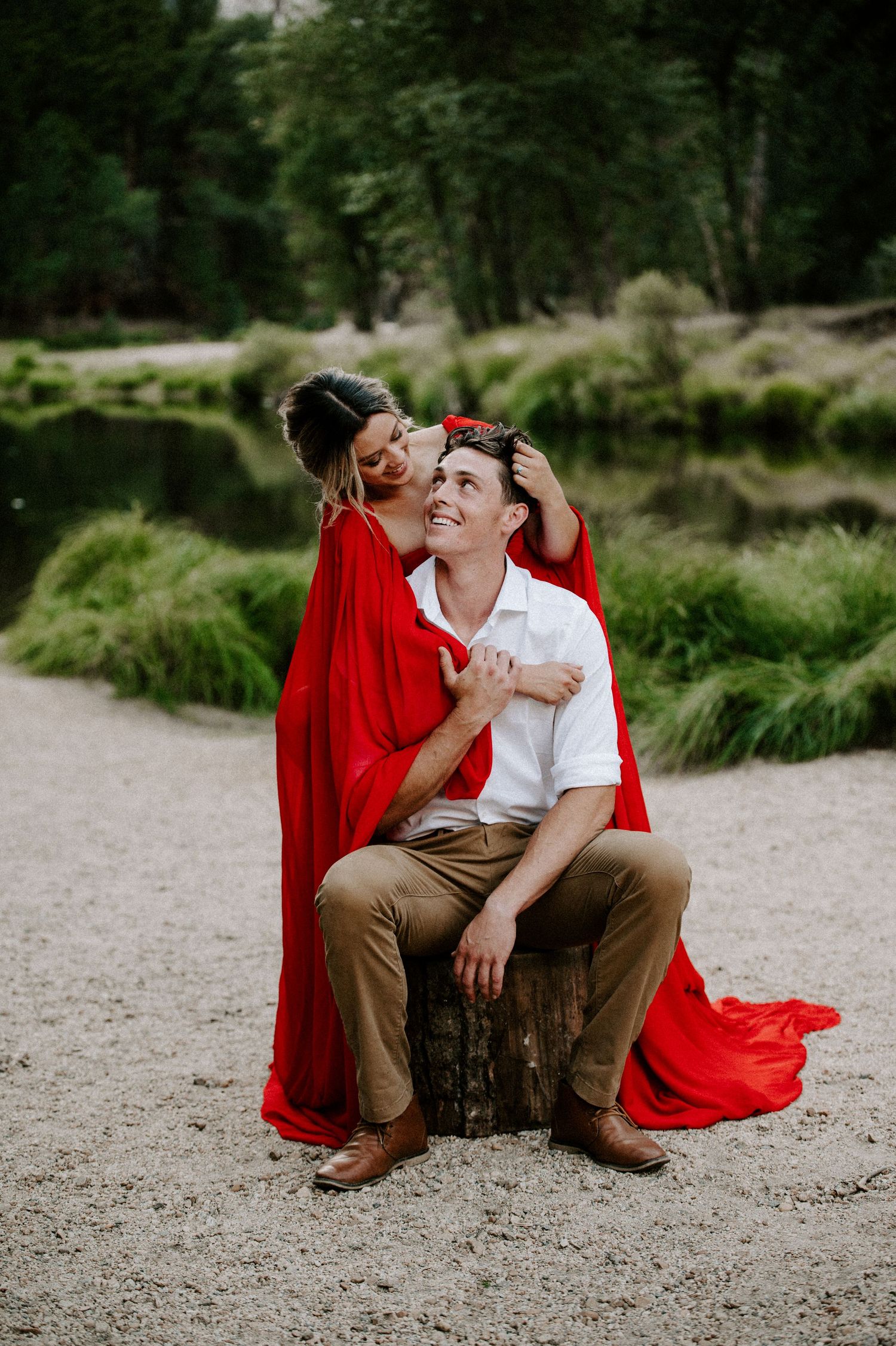 A couple shares an intimate moment on a tree stump in a lush park setting with a red blanket.